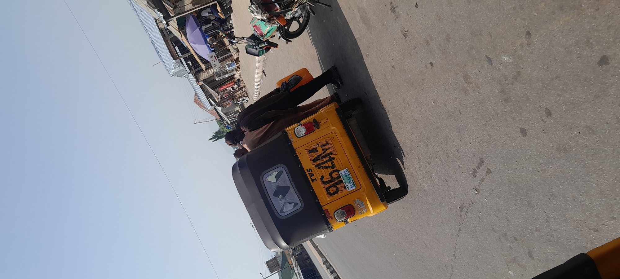 The image shows a street scene in Jalingo, Nigeria, with a yellow auto-rickshaw in the foreground. A person is seated in the auto-rickshaw, wearing a dark head covering and dark clothing. The auto-rickshaw has a license plate with the text "KLB-679DU" and "NIGERIA." There are other vehicles, including a motorcycle, and what appears to be market stalls in the background. The scene suggests a bustling street environment, likely during daytime given the bright lighting and shadows. The sky is clear and blue, indicating fair weather. The overall impression is of a typical day in a Nigerian town, with local transportation and commerce in full swing.