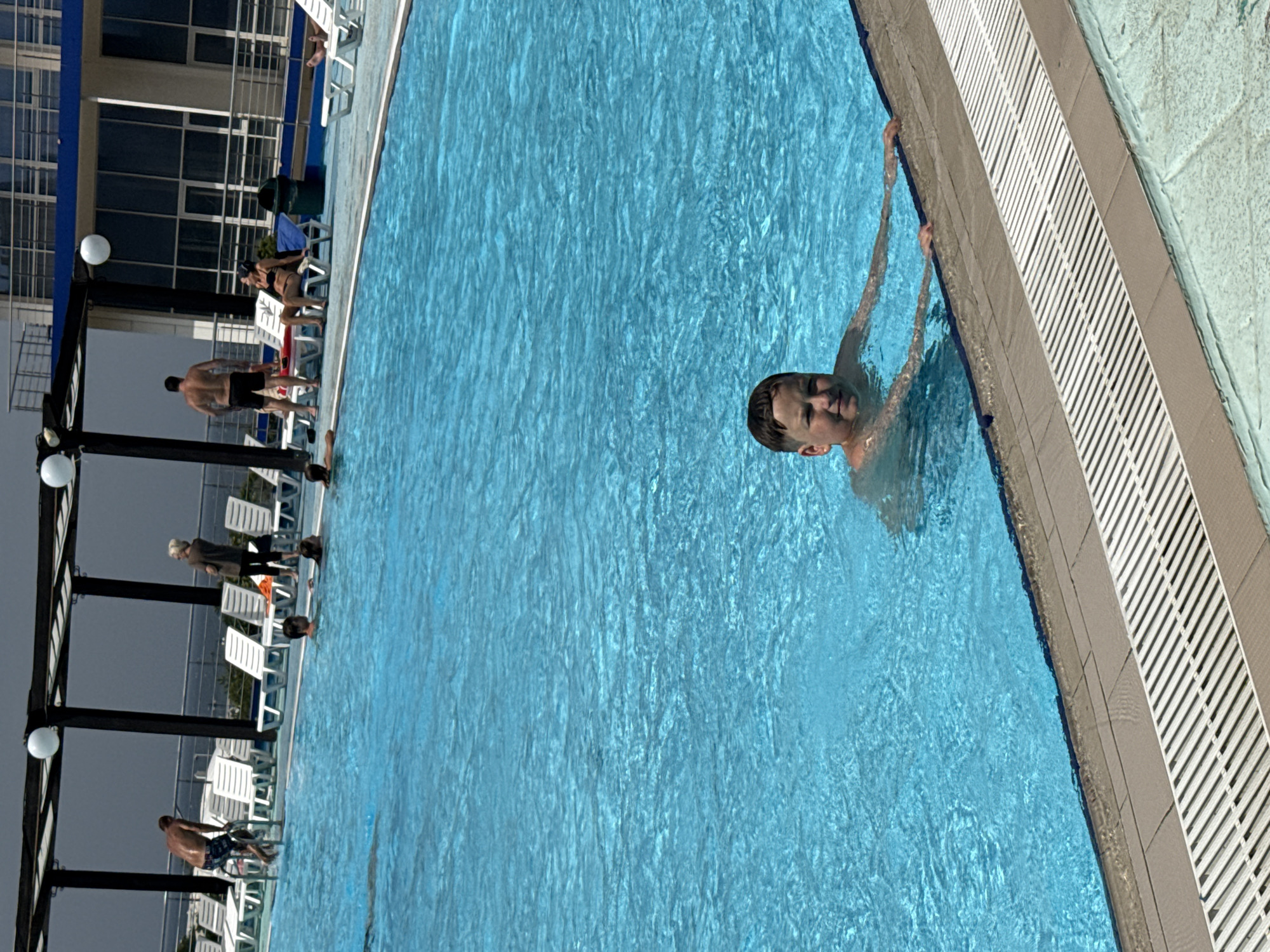 The image shows a boy swimming in a pool at a seaside resort, likely the "President Hotel" in Sevastopol, Ukraine, given its distinctive architecture and seaside location. The pool is large and blue, with clear water. Several people are visible around the pool, lounging on chairs or swimming. The boy in the foreground is looking at the camera with a smile. The background features the hotel building and a cloudy sky. The overall atmosphere is one of leisure and relaxation.