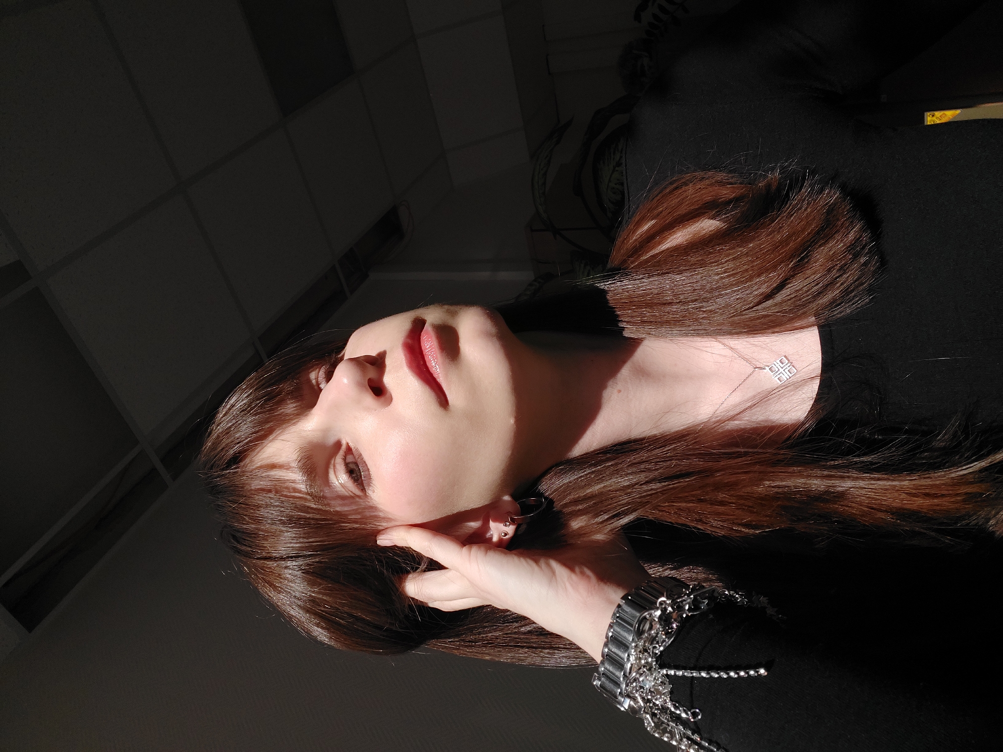 A low-angle, indoor portrait features a woman with long brown hair and fair skin, her face partially illuminated by a strong light source from the upper left. The light casts sharp shadows, emphasizing the ceiling tiles in the background. She is wearing a black top, a silver necklace with a square pendant, and a large, multi-strand silver bracelet on her left wrist. Her left hand is near her face, fingers gently touching her cheek. The composition and lighting create a dramatic effect. There is no indication or evidence of this picture being taken in Chaikovsky, Russia.