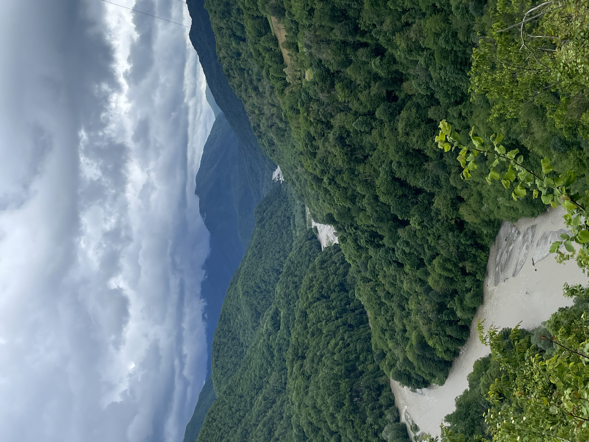 The image captures a mountainous landscape under a cloudy sky. A river flows through a valley densely covered with green trees. The mountains recede into the distance with varying shades of green. The sky is dominated by gray clouds. The image shows no evidence of people, subjects, or objects. The location is not evident from the image itself, so there is no indication of the city of Khartsyz Pervyi, Russia.