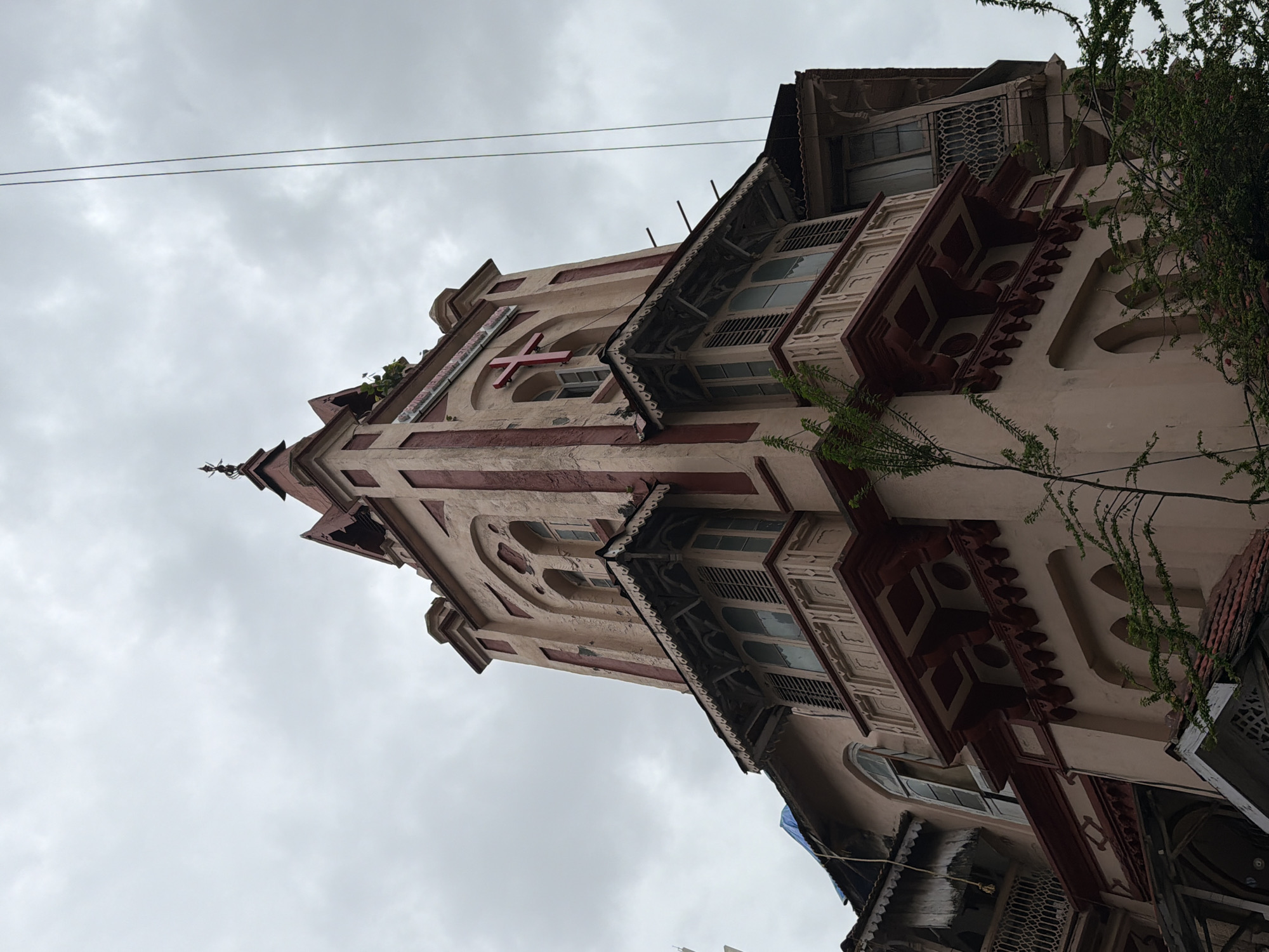 The image shows a close-up, low-angle view of the exterior of a building with a prominent tower, located in Mumbai, India. The architecture appears to be colonial era, with a light pink facade accented by dark red trim and decorative elements. The building features multiple levels with arched windows, balconies with intricate wooden detailing and latticework, and a red cross mounted on the upper section of the tower. The sky is overcast and grey. A single strand of electrical wire is visible extending down from the top right of the frame. Lush green foliage partially obscures the lower sections of the building.
