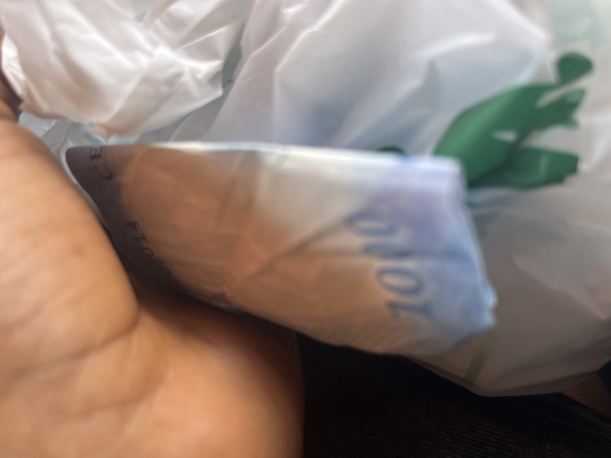 In the city of Gura Top, Nigeria, an image captures a close-up of a person's hand holding a wad of Nigerian Naira currency, specifically a 1000 Naira note. The banknote displays the number "1000" in large blue digits. The currency appears slightly creased and is partially enclosed within a white plastic bag. A portion of a green object is visible within the bag, though its exact nature is unclear due to the blurriness. The background is out of focus, with hints of other objects and the general ambiance of an indoor or covered setting. The lighting suggests it is daytime. The image composition is tightly framed, focusing on the currency and the hand.
