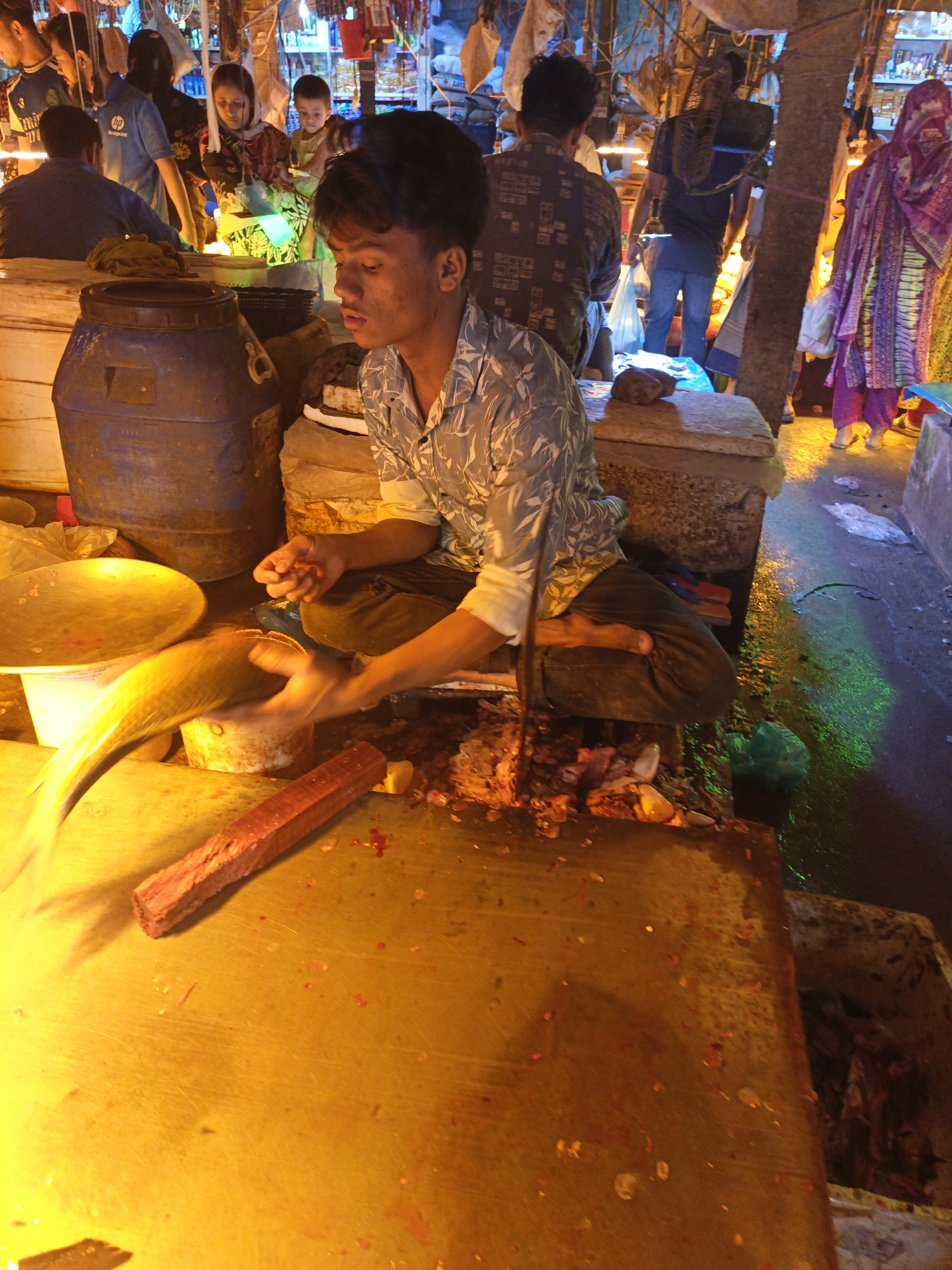 A fishmonger sits on a concrete ledge in Dhaka, Bangladesh, preparing fish at a market stall. He wears a patterned gray shirt and dark pants, seated cross-legged. A large yellow fish is being prepared on a yellow-toned table covered with remnants of fish processing. A wooden block and bowl are nearby. Behind him are barrels and customers shopping at adjacent stalls. The environment appears to be a bustling, wet market under bright, somewhat harsh lighting. The ground is damp, with reflections, and discarded fish parts are visible.