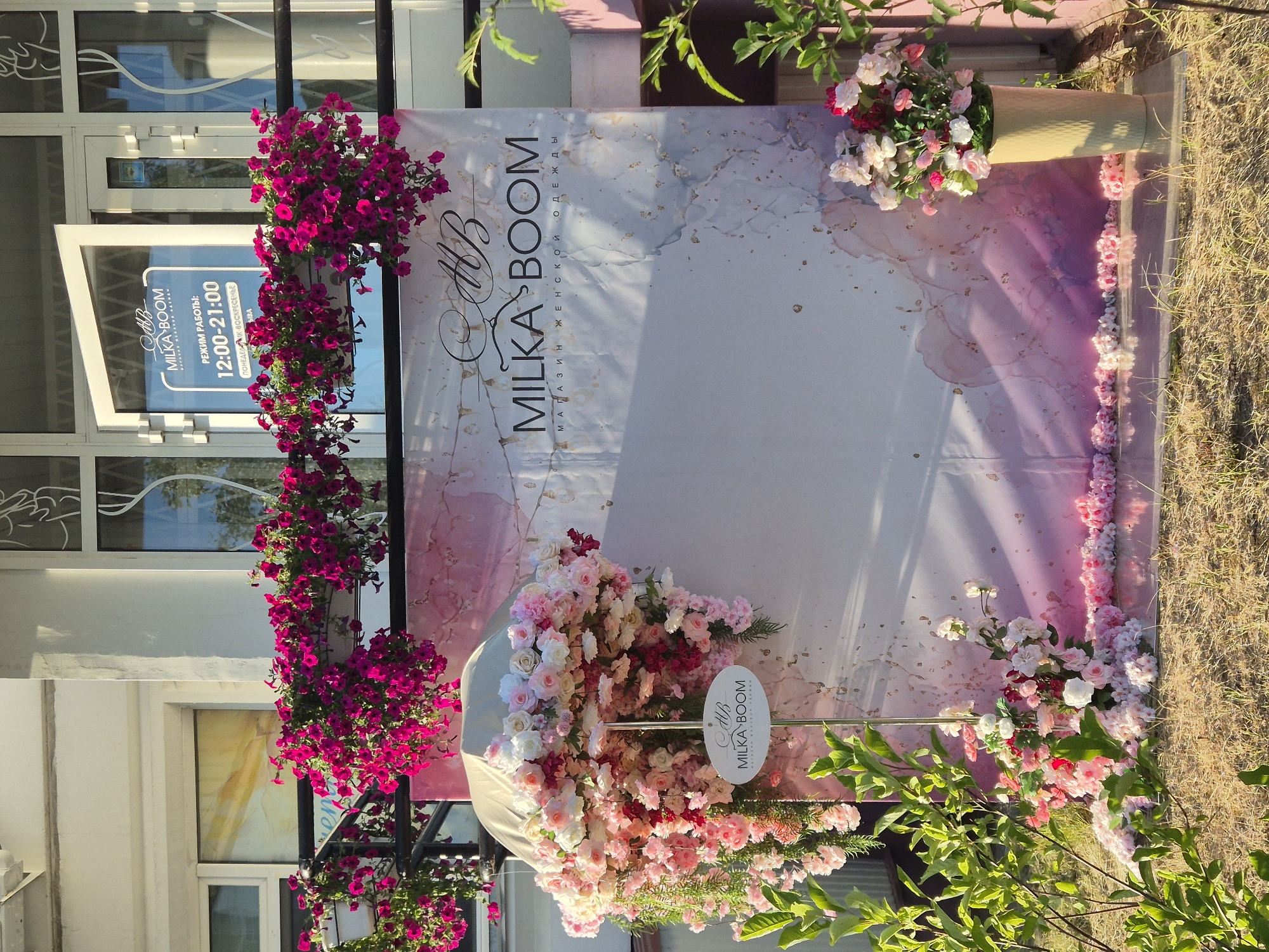 This is a vertical, outdoor photograph of a storefront decorated with flowers and a banner. The storefront, on the left side of the image, has large windows and a white exterior. A sign on one of the windows indicates the business name "MILKA BOOM" and lists its operating hours as 12:00-21:00 daily. The banner behind the flowers features a marble-like pattern in shades of pink and white with gold accents. The text on the banner also reads "MILKA BOOM" and "МАГАЗИН ЖЕНСКОЙ ОДЕЖДЫ," which translates to "WOMEN'S CLOTHING STORE."
Cascading arrangements of bright pink petunias hang from planters to the left of the banner, and a large, lush bouquet of pink and white roses is placed at the base of the banner, partially covering a white elliptical sign with the "MILKA BOOM" logo. More pink flowers are arranged in a line along the ground in front of the banner, and a tall, light-yellow vase filled with similar pink and white flowers stands to the right of the banner. Green foliage is visible in the foreground and upper right corner, suggesting an outdoor setting bathed in sunlight, likely in the late afternoon or early evening, given the warm lighting and long shadows. The overall impression is one of a festive and welcoming storefront display.