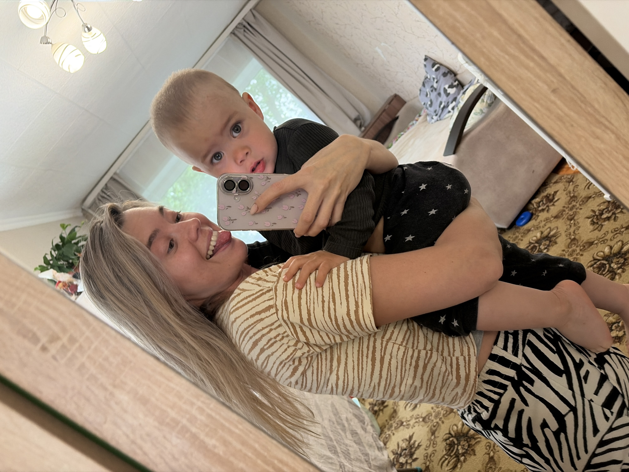 A mirror selfie captures a woman holding a baby in what appears to be an indoor home setting. The woman, with long blonde hair, is smiling broadly with her tongue playfully extended, and is wearing a cream-colored top with brown vertical stripes and black and white zebra-print pants. She is holding a light pink smartphone with a floral pattern case, which is being used to take the selfie. The baby, with short light brown hair and blue eyes, is held by the woman and looks directly into the phone's camera with a curious expression. The baby wears a dark ribbed long-sleeve top and black pants with white star patterns.
The reflection shows parts of a room, including a white ceiling with a modern light fixture, a window with light-colored curtains, and a bed or sofa covered with a patterned blanket or rug. The lighting suggests it is daytime. The image is taken at an angle, showing the wooden frame of the mirror in the foreground and background. Both subjects appear happy and engaged in the moment. No discernible text is visible on clothing or objects.