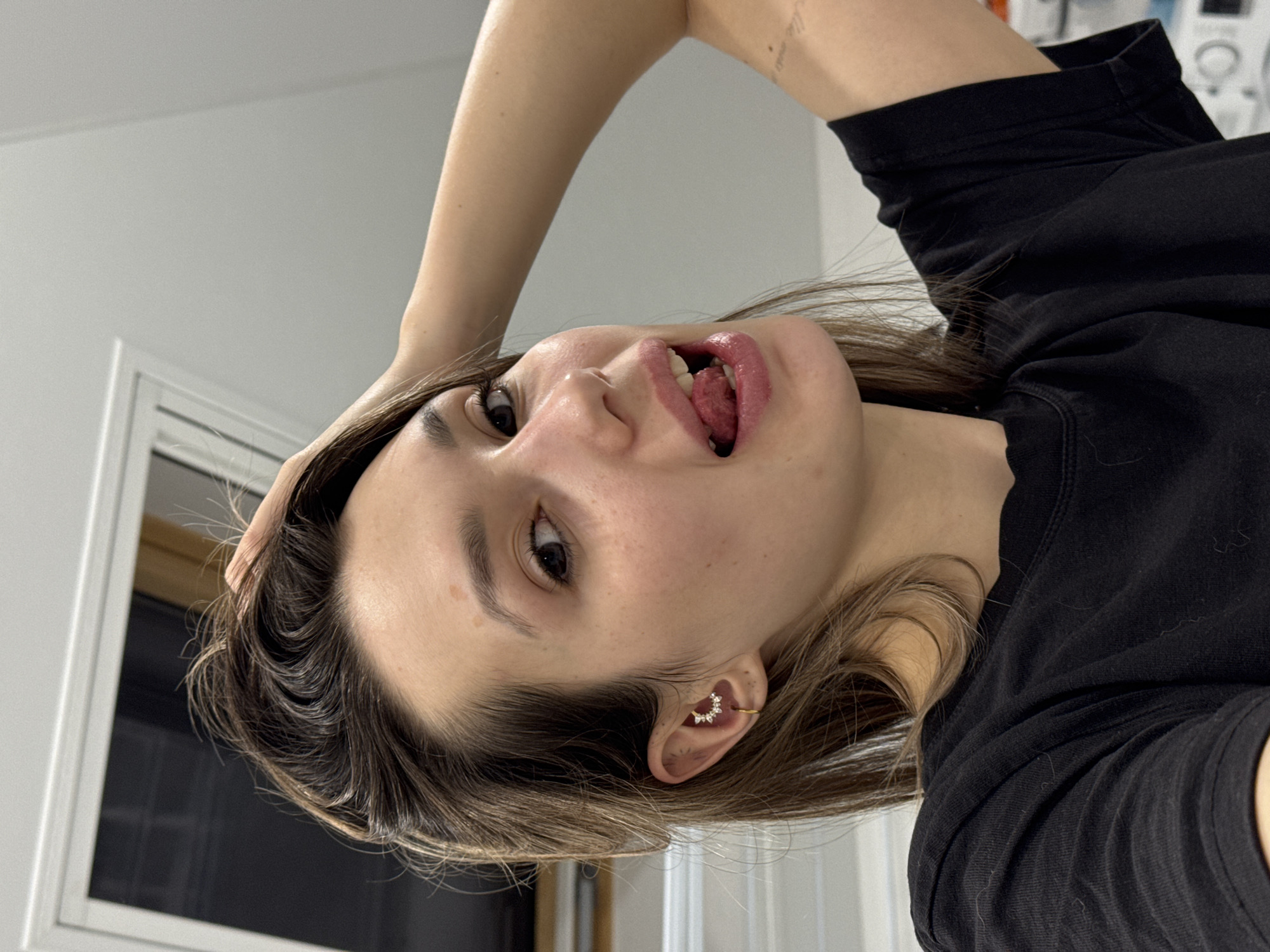 A woman with brown hair and fair skin, wearing a black t-shirt, is posing with her tongue sticking out. She is holding the side of her head with her hand, and her right ear is adorned with a gold-colored ear cuff. The background shows a white wall and a window frame. The location is Unknown, Unknown.