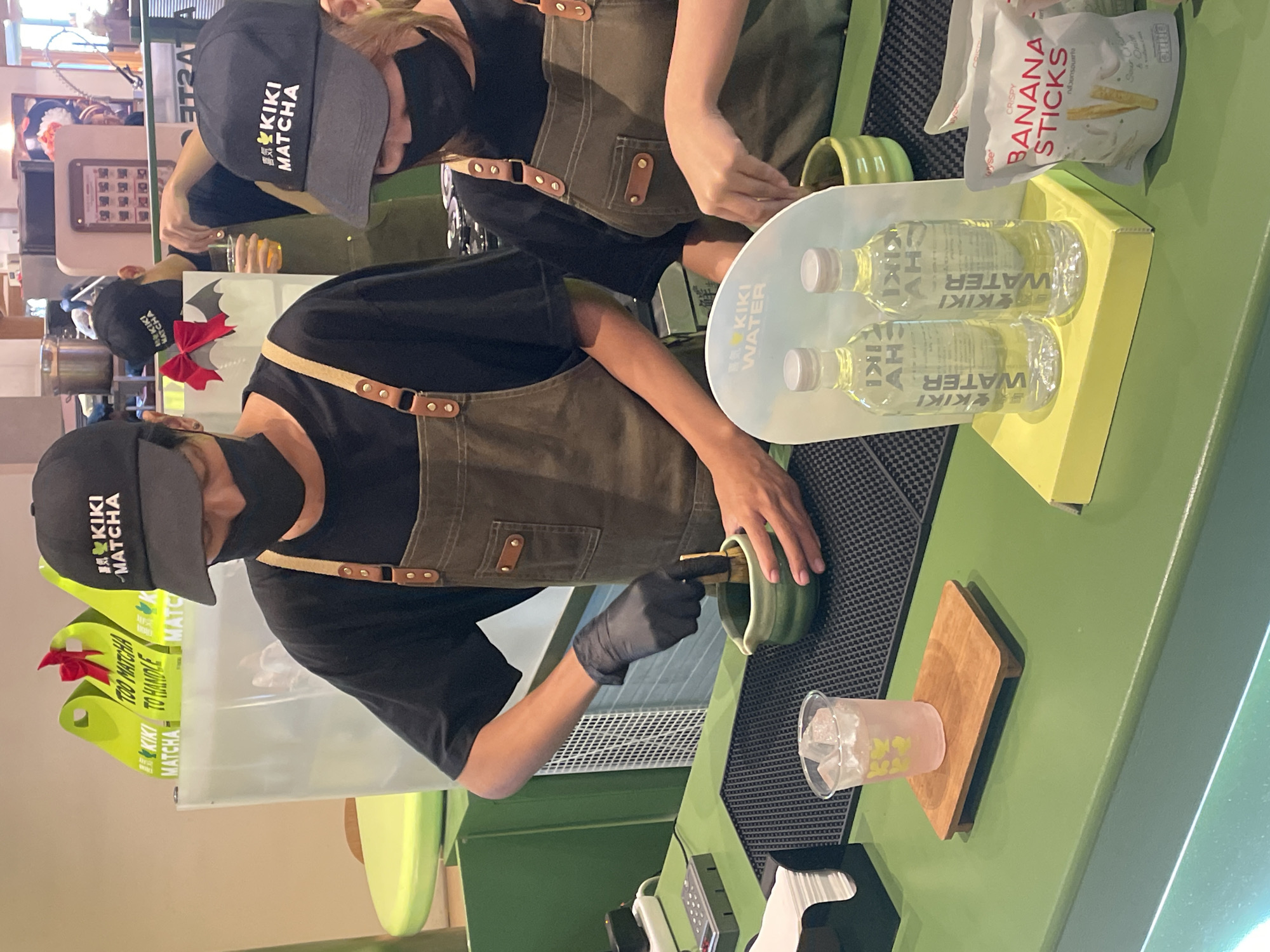 The image shows the interior of a Kiki Matcha establishment, likely a tea shop. Two employees wearing black baseball caps with the "Kiki Matcha" logo, black face masks, and olive-green aprons are working behind a green counter. The apron has leather straps with a metal clasp. One employee, wearing black gloves, is using a whisk to prepare matcha in a green cup. Nearby is a plastic cup filled with ice on top of a wooden square and two bottles of Kiki water inside a white plastic holder. In the background, there is a bag of Crispy Banana Sticks. The image suggests a close-up view inside the shop. Given the brand name, Kiki Matcha, and the presence of the Thai word "กล้วยกรอบ," which means "crispy banana" the location is likely Bangkok, Thailand.
