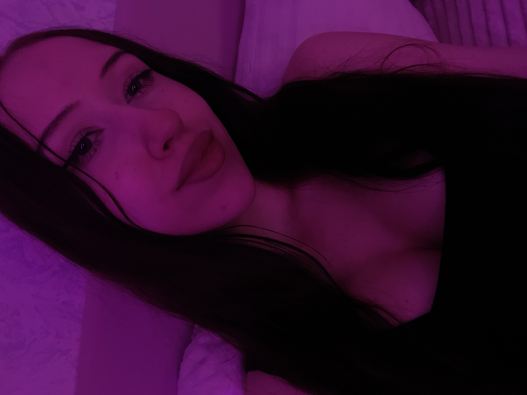 A young woman with long dark hair is captured in a close-up, selfie-style shot. She is wearing a black top and is bathed in a pinkish-purple light. The background appears to be a textured wall and a pillow, suggesting an indoor setting, possibly a bedroom. The lighting creates a soft, atmospheric mood. The image does not contain any information that would allow for the determination of its location in Votkinsk, Russia.