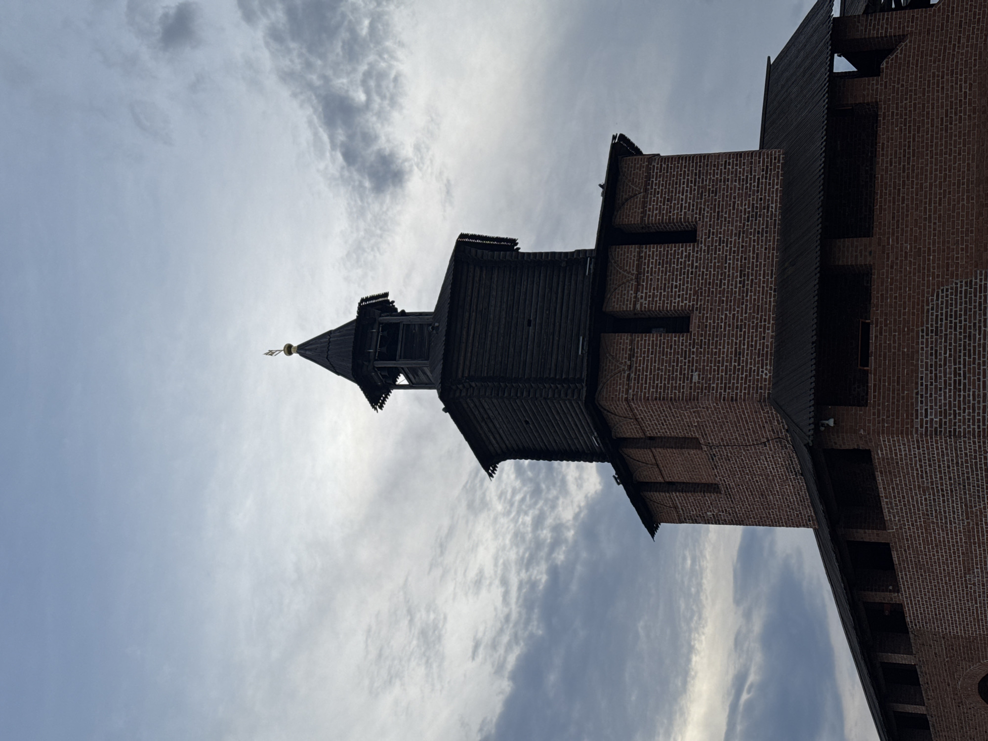 The image captures a section of the Nizhny Novgorod Kremlin in Russia, specifically a tower structure. The architectural elements include a wooden conical roof on top of a brick tower. The tower is constructed of red bricks and has several narrow openings or windows. The wooden portion is dark in color and appears to be aged. The sky is overcast with a mix of light and dark clouds, creating a backdrop to the structure. There are no people visible in the frame. The low angle of the shot emphasizes the height of the tower.