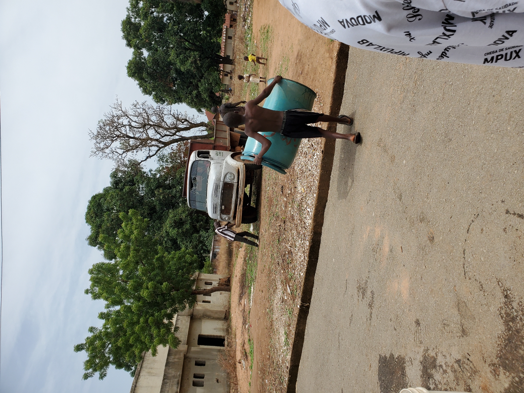 A young, shirtless boy in black shorts struggles to lift a large, light blue barrel, likely full of water, onto the back of a white truck parked along the side of a paved road. Another person stands nearby near the front of the truck, while two smaller children play in the dirt area behind the truck. A concrete building with open windows sits to the left of the frame, partially obscured by trees. Overhead, part of a shirt with printing on it is visible. The background shows a mixture of dirt, grass and vegetation, suggesting a partly developed or less affluent area of Mararaba, Nigeria.