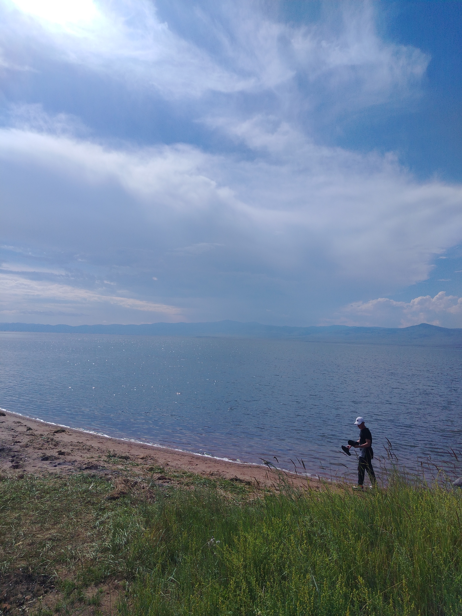 A man stands on the sandy shore of a large body of water, holding a pair of black shoes in his left hand. He is wearing a white baseball cap, a dark t-shirt, and dark pants. The water, which appears to be a lake, is calm with a slight ripple and reflects sunlight. In the distance, a range of hazy, blue mountains stretches across the horizon under a partly cloudy sky. The foreground consists of tall, green grass and a sandy beach. The scene is likely located in Tsaydam, Russia, given the geographical context of large lakes and mountainous horizons.