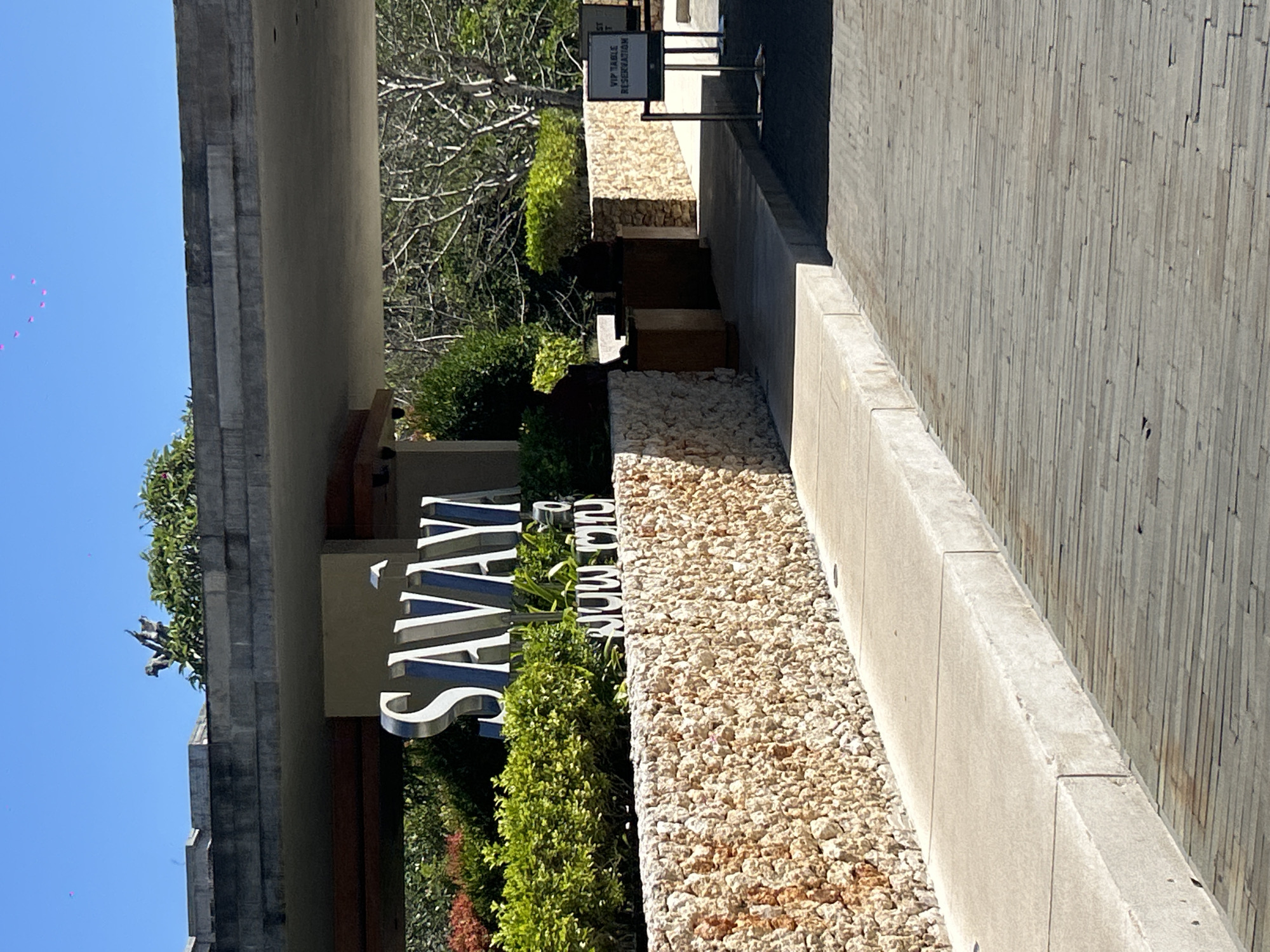 The image shows the entrance to the Savaya Bali resort in Kuta Selatan, Indonesia. A large, silver "SAVAYA" sign is mounted on a stone wall. In front of the sign, a pathway leads to the resort's entrance, which is partially obscured by greenery and trees. A sign indicating "VIP TABLE RESERVATION" is visible to the right of the pathway. The sky is clear and blue, and the sun is shining. The resort appears to be nestled in a tropical setting with lush vegetation and stone architecture.