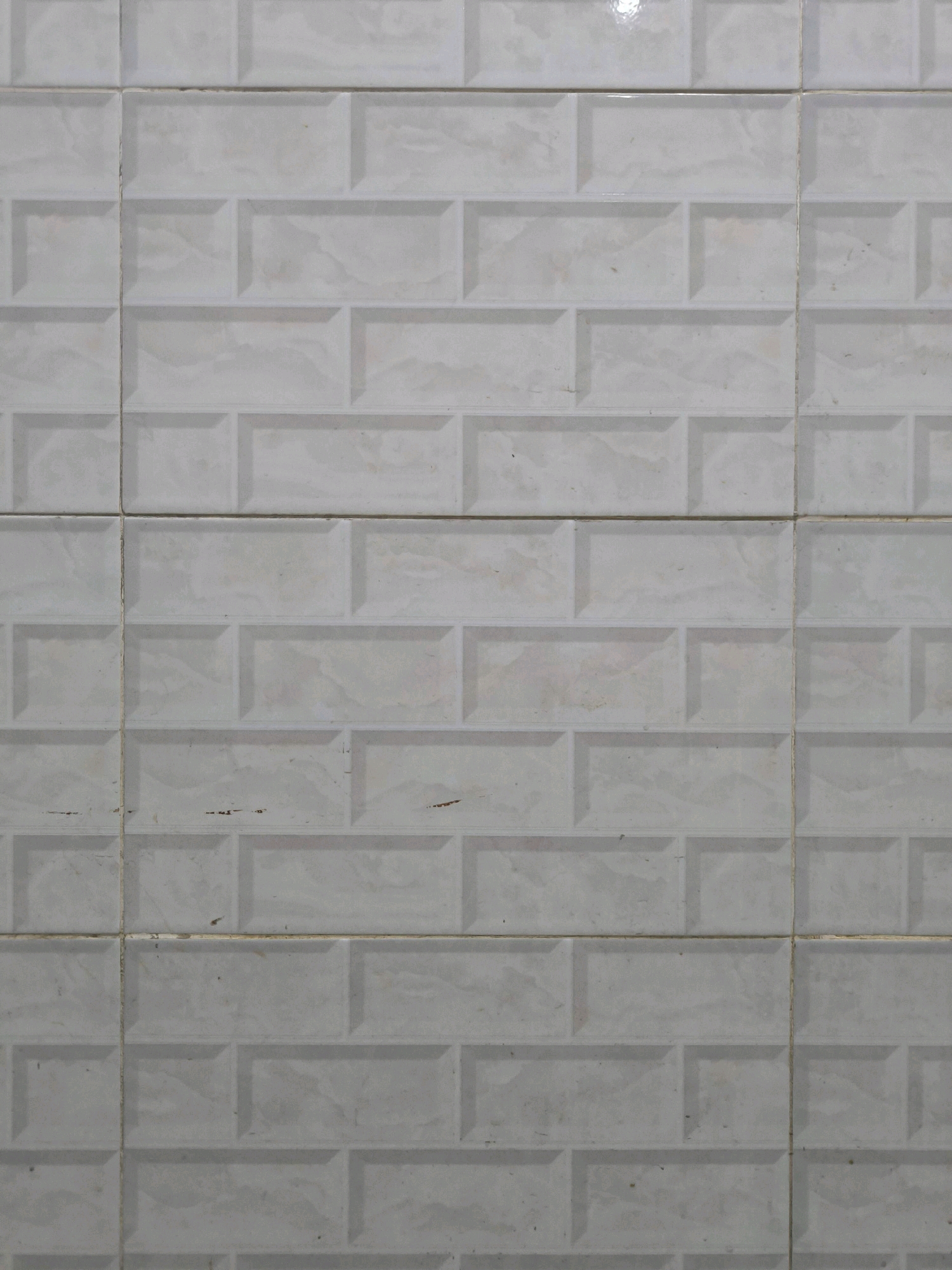 A wall is covered in off-white, rectangular tiles arranged in a running bond pattern. The tiles have a subtle, marble-like texture with faint gray veining. Each tile has a raised, beveled edge, creating a dimensional brick-like appearance. The grout lines between the tiles are a light yellow color and appear slightly uneven. There are a few small, dark marks and some dirt smudges on the tiles, particularly in the lower half of the image. The lighting suggests a diffused, indoor environment. The image does not depict any people, objects, or actions. This scene is a close-up of a tiled wall, likely found in a bathroom or kitchen, and its location in Tabanan, Indonesia, is not visually evident from the image itself.