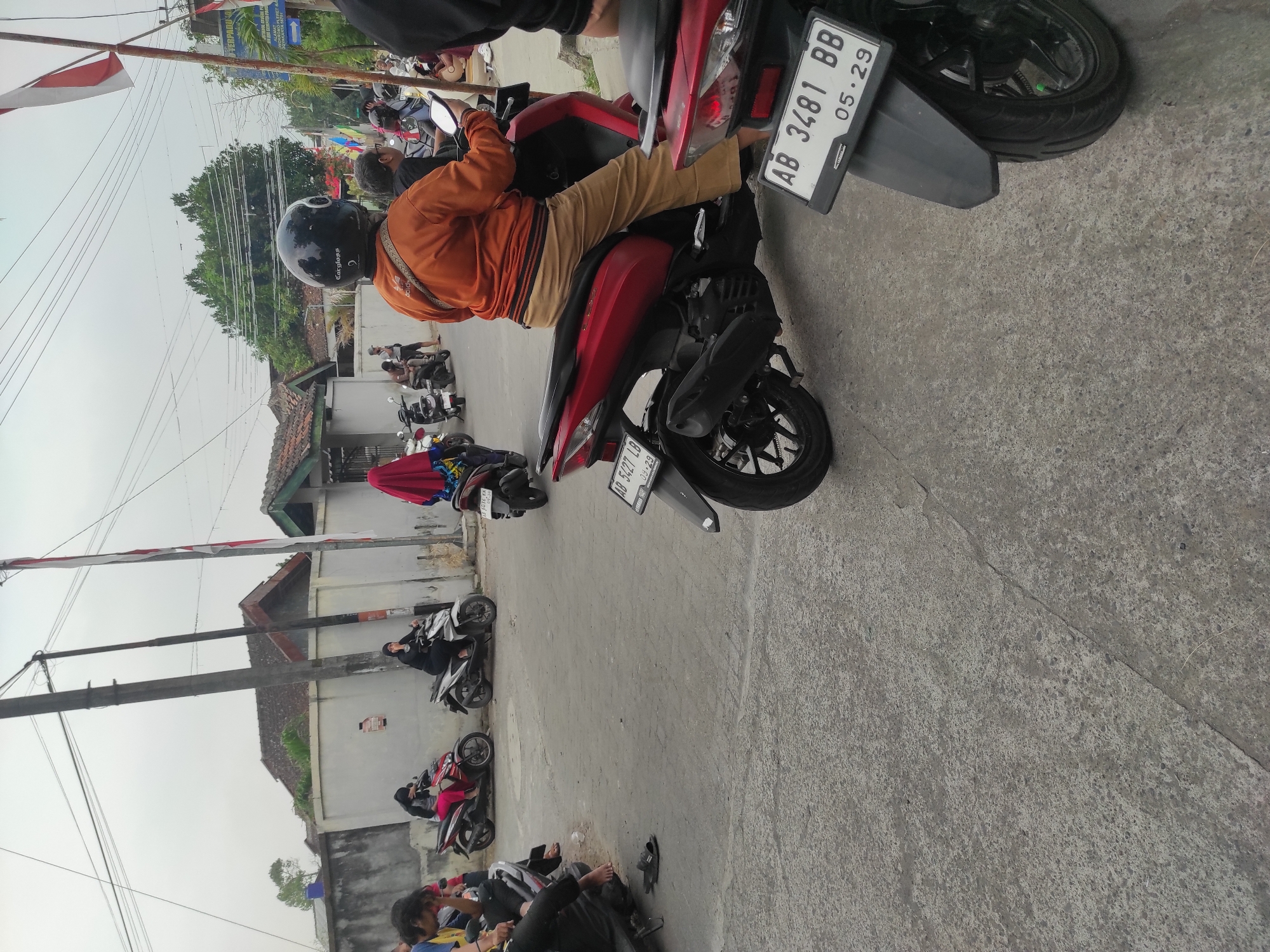 In Pleret, Indonesia, a street scene unfolds with multiple motorcycles parked and in motion. A person wearing an orange long-sleeved shirt and tan pants, along with a black helmet, rides a red scooter. Behind them, another red scooter with a license plate reading "AB 3481 BB 05.29" is visible. Further back, several other individuals on motorcycles are present, some in motion and others stationary. The environment appears to be a road or parking area with buildings and electrical wires overhead. The sky is overcast.