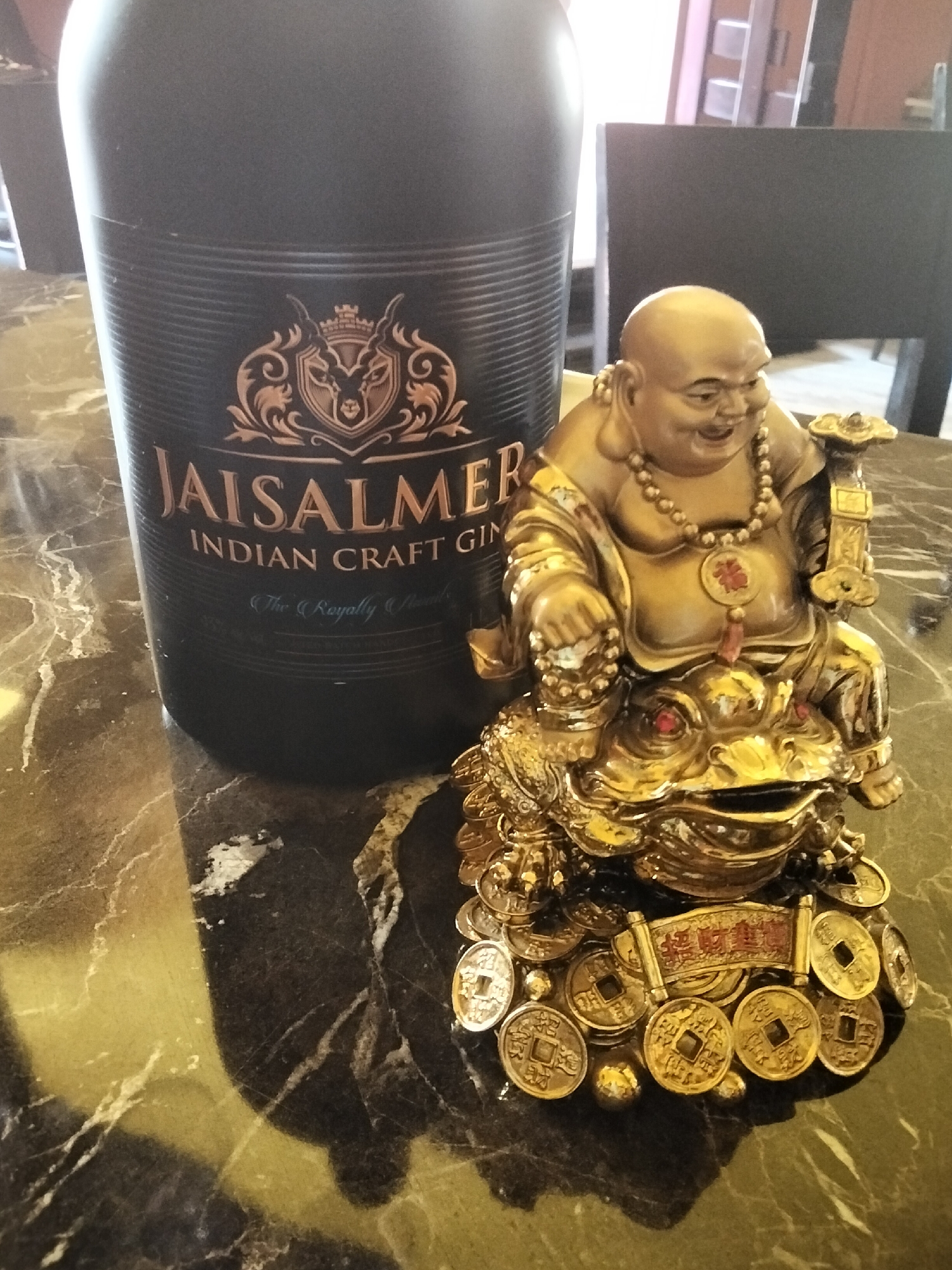 An indoor, medium shot captures a bottle of Jaisalmer Indian Craft Gin and a golden statue of a laughing Buddha seated on a toad and surrounded by coins. The bottle, a dark matte black with gold lettering and detailing, stands to the left of the frame. The statue, a bright gold, is positioned to the right and slightly in front of the bottle. The Buddha is depicted with a jovial expression, wearing a beaded necklace and holding a scepter. The toad it sits on is adorned with red gemstones, and the base is covered in gold coins, some of which display Chinese characters. The objects rest on a dark, polished surface with a marbled pattern, likely a table. The background is blurred, showing muted tones of furniture and walls, suggestive of an indoor domestic setting. There are no people present in the image.