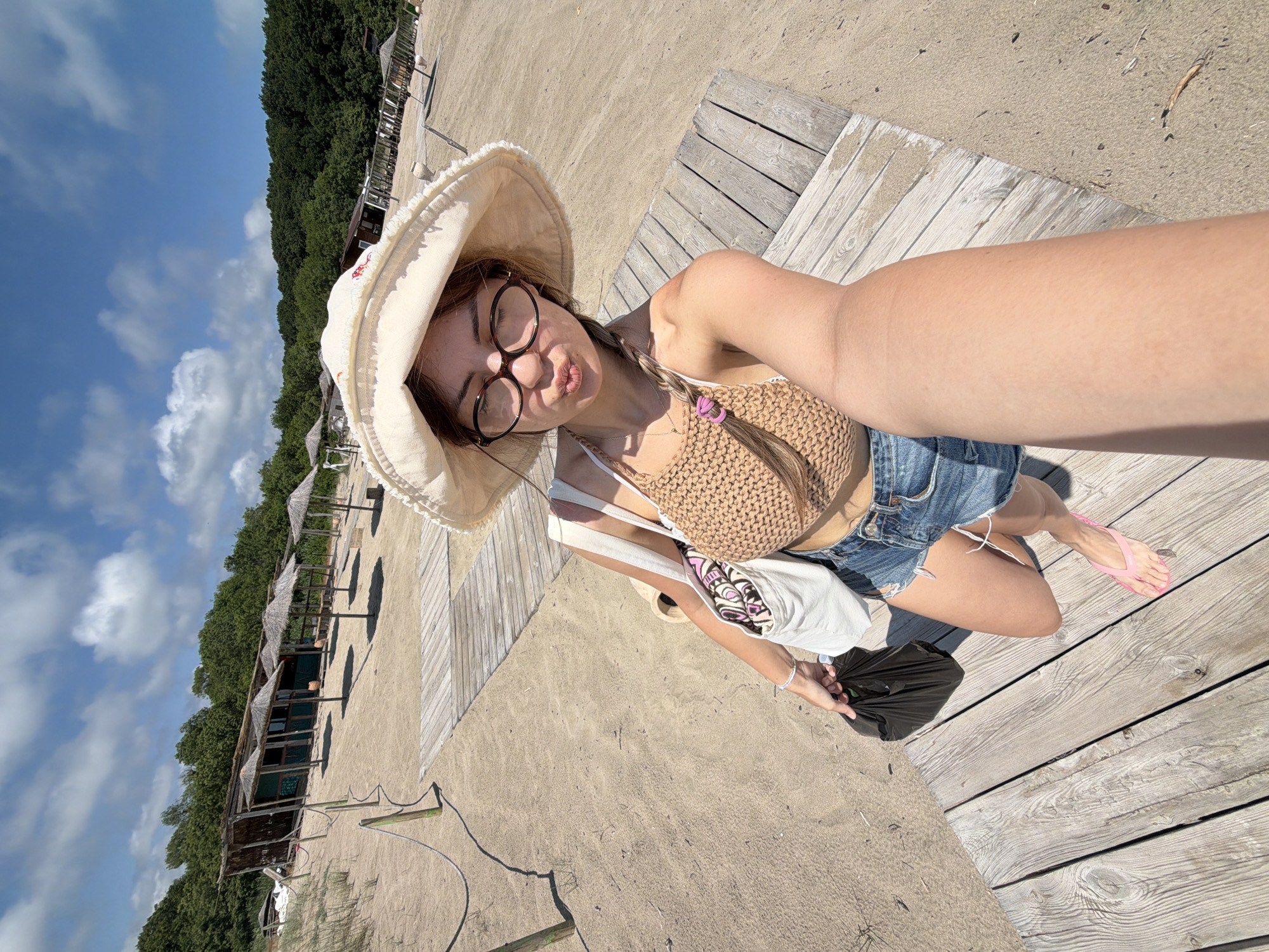 The image depicts a young woman on a sandy beach, likely in Rezovo, Bulgaria, given the context of the provided location. She is wearing a wide-brimmed hat, round glasses, and a crocheted halter top with denim shorts. Her right arm is extended, holding a black plastic bag. She is standing on a wooden boardwalk, which appears to be a pathway on the beach. In the background, there are thatched-roof beach umbrellas and some small wooden structures, suggesting a resort or beachside establishment. The sky is blue with scattered white clouds, and a line of trees forms the horizon behind the beach structures. The woman is posing for a selfie, with her lips pursed. She is also carrying a white tote bag with a patterned scarf peeking out. A pink flip-flop is visible on her left foot. The overall scene suggests a sunny day at the beach.