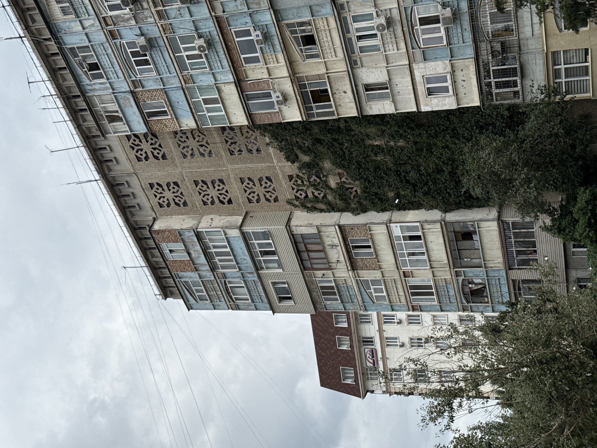 An overcast day in a city shows a multi-story apartment building with a pale blue and beige facade. The building has many windows, some of which are closed, and others open with balconies. Air conditioning units are visible on several floors. The building is partially covered in a thick layer of ivy on its left side. To the left of this building is another apartment building with a white facade and a dark brown pitched roof with several dormer windows. In the foreground, lush green trees partially obscure the lower floors of the buildings. Overhead, grey clouds fill the sky, and power lines are strung across the scene.