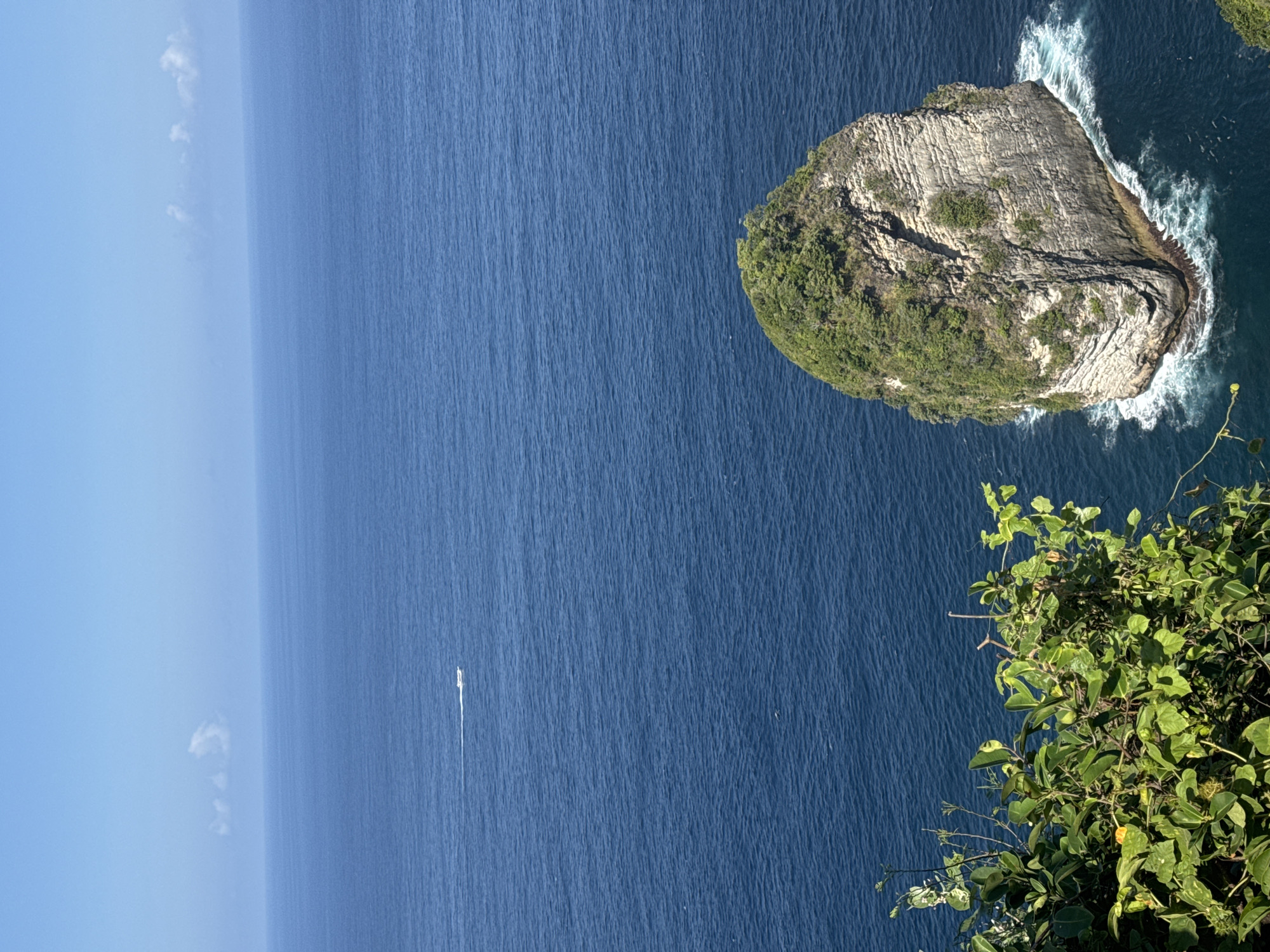 The image depicts a stunning seascape in Nusa Penida, Indonesia. In the foreground, lush green foliage cascades down from a cliff, framing a breathtaking view of the ocean. The deep blue water stretches to the horizon, interrupted by a small white boat leaving a wake. A prominent rock formation, covered in dense green vegetation, rises majestically from the sea, its base being pounded by white-capped waves. The sky above is a clear, vibrant blue, with a few wispy clouds dotting the upper part of the frame. The scene conveys a sense of natural beauty and tranquility.