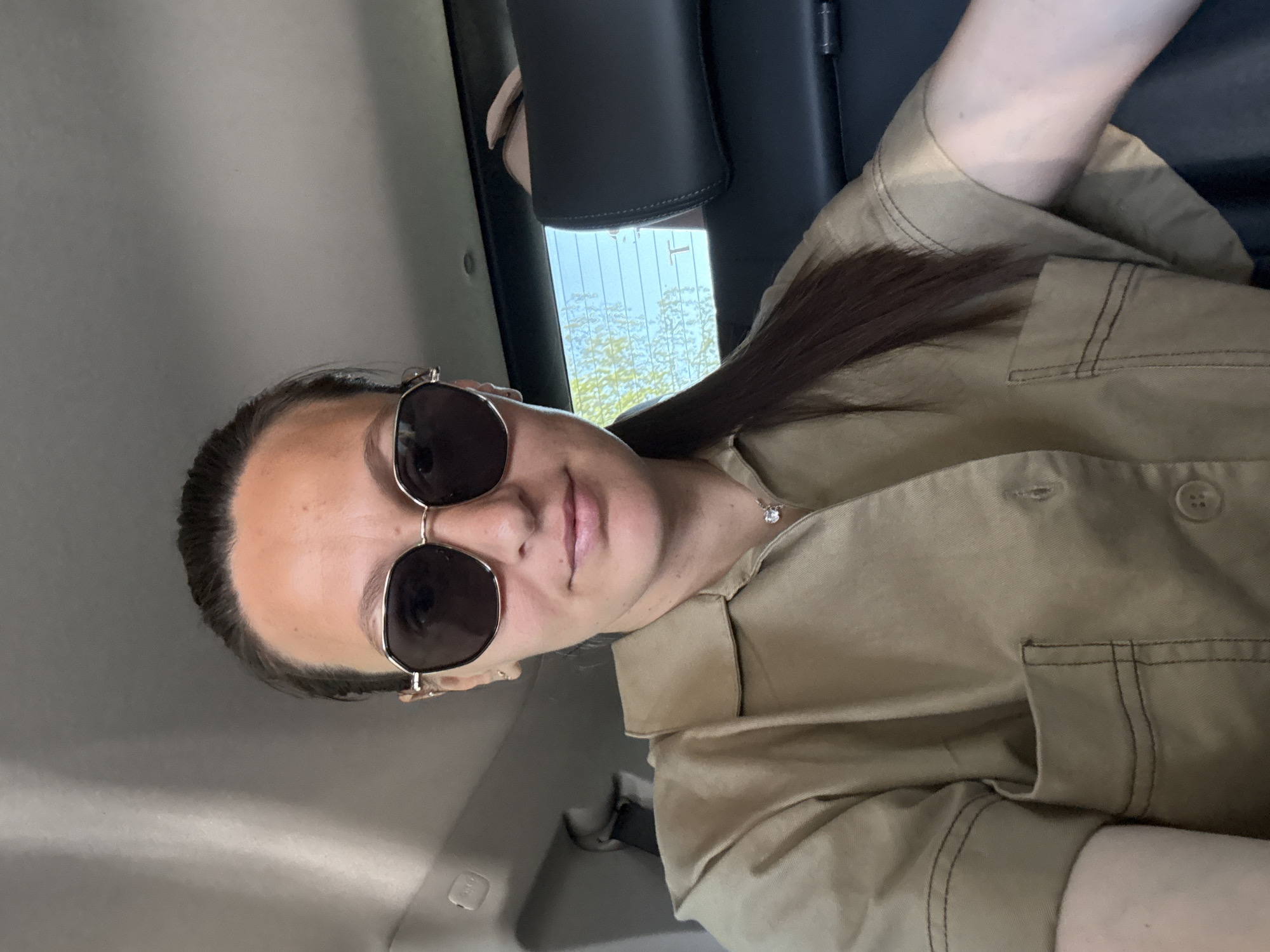 The image is a selfie taken from a low angle inside a vehicle. A person with dark hair, wearing sunglasses and a beige shirt, is the main subject. A glimpse of the outside through the car window reveals trees and possibly a sky. The background indicates the interior of the vehicle, including a headrest. There's no specific information to suggest the photo was taken in Chaikovsky, Russia, or any city location.