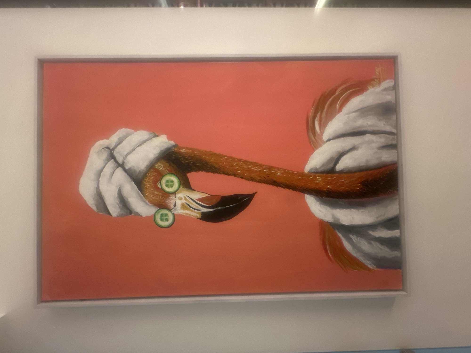 A framed painting depicts a flamingo receiving a spa treatment. The flamingo has a white towel wrapped around its neck and head, with cucumber slices on its eyes. The background of the painting is a flat coral color. The location is North Kuta, Indonesia.