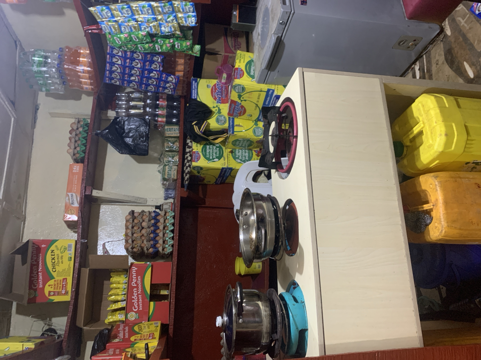 This image shows the interior of a small convenience store or stall, likely in Gura Top, Nigeria. The shelves are packed with various goods including instant noodles, bottled drinks, eggs, and what appear to be large containers of water or other liquids. On a counter in the foreground, two cooking pots are placed on portable stoves. One pot is larger and stainless steel, while the other is a wider, shallower stainless steel pot. Behind the counter, there is a large grey refrigerator and additional shelving with more products. The overall environment appears to be a working retail space with limited lighting.