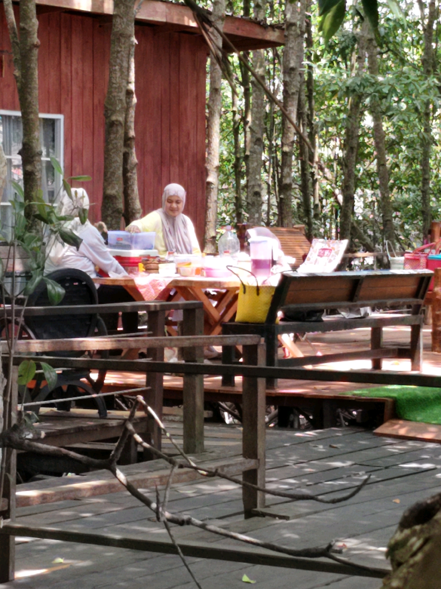 An outdoor scene depicts two individuals seated at a table in front of a wooden building. The building has red plank siding and a window. To the left, a person in a white shirt is partially visible. To the right, a person wearing a yellow shirt and a light purple headscarf is seated at the table, appearing to interact with items on it. The table is laden with various containers, including plastic tubs and bowls, and a checkered red and white cloth. A yellow bag rests on the table. In the foreground, a wooden deck and railing are visible, with tree trunks and lush green foliage surrounding the area. The scene suggests a casual outdoor dining or gathering setting, possibly in a restaurant or resort, given the environment and setup. The lighting indicates it is daytime, with shadows cast across the deck.