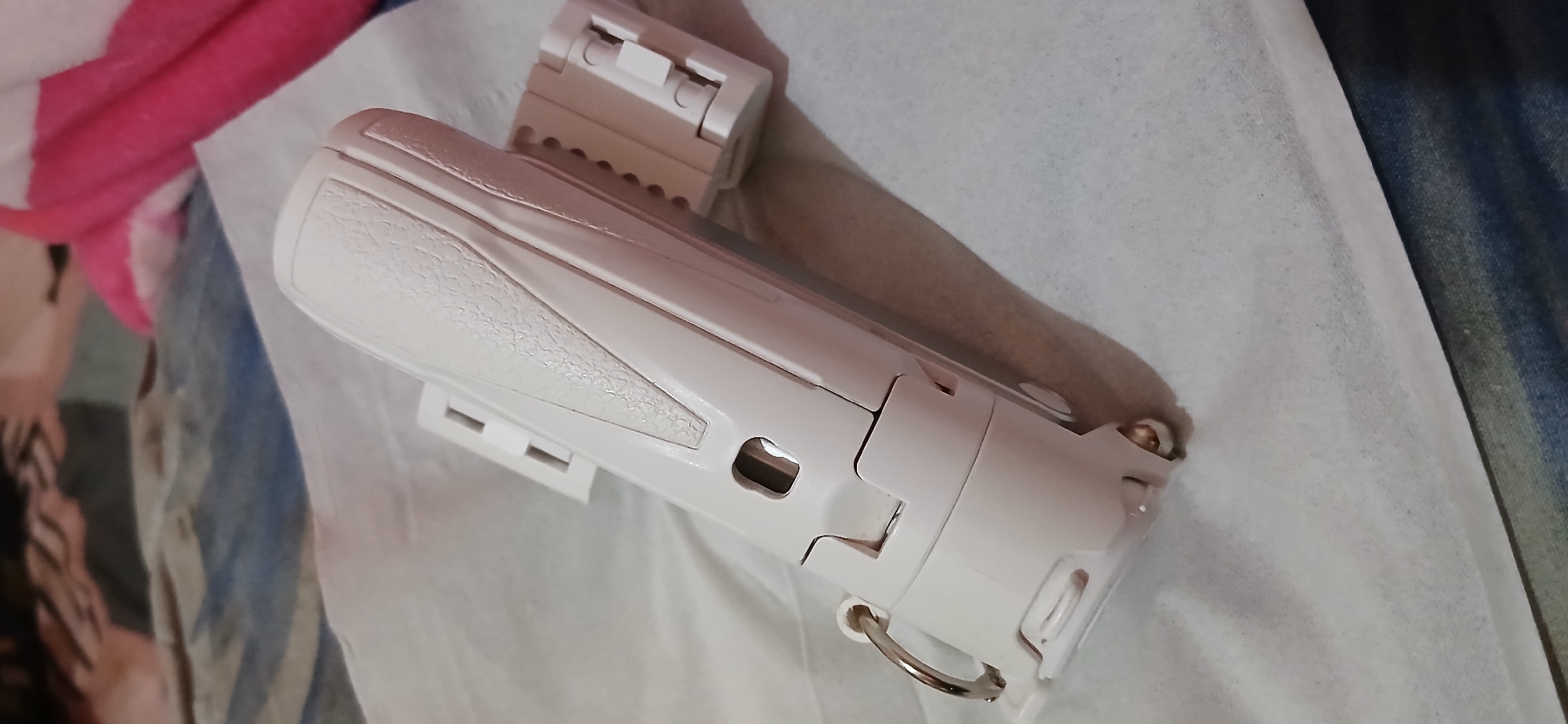 The image displays a white electric beard trimmer with a textured grip. The trimmer is lying on a white textured surface, possibly paper or cloth. Two white attachment pieces are connected to the trimmer, one on each side, suggesting it's a disassembled part of a grooming kit. In the background, out of focus, there appears to be bedding with blue and white striped patterns and a pink fuzzy fabric. The scene is indoors, and the lighting suggests it's daytime. There are no people visible in the image. The location in Samarinda, Indonesia, cannot be determined from the visual information provided.