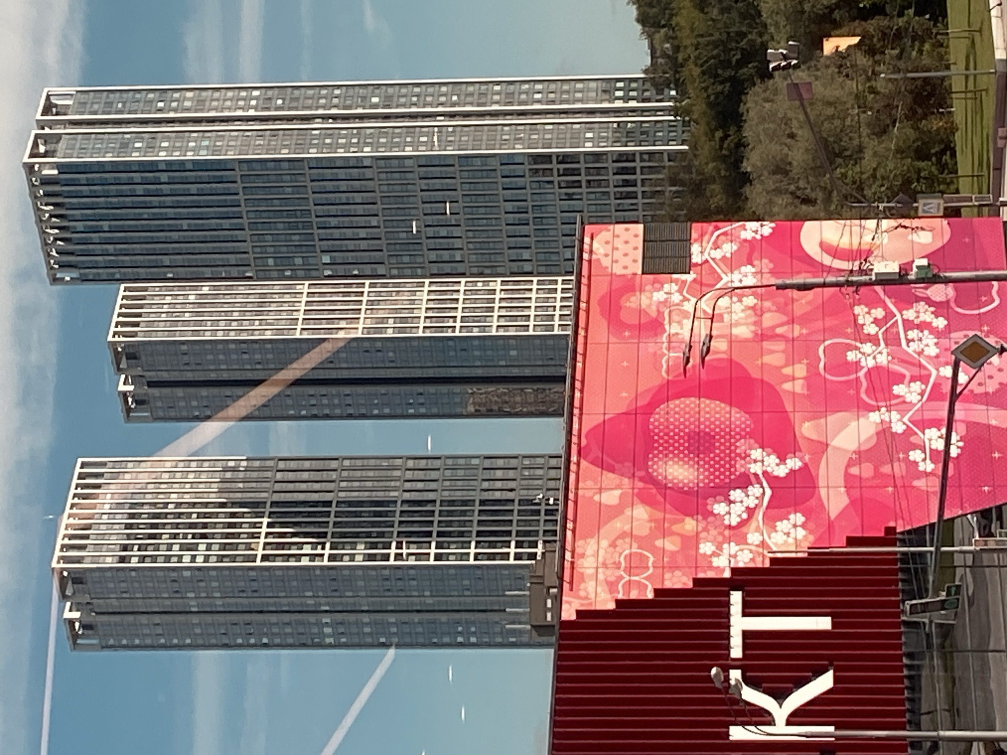 The image shows a close-up of three modern skyscrapers under a clear blue sky with a few faint clouds. The skyscrapers are tall and slender, with numerous windows. The building on the left is lighter in color than the other two, which have a darker, glass facade. In the foreground, a large, brightly colored mural is visible on a building with a red, ribbed facade. The mural is predominantly pink and features floral patterns and heart shapes. The letters "KT" are visible in white on the red portion of the facade on the left. A street lamp is positioned in front of the mural. The background shows a lush green hillside with trees, and a few streetlights and traffic signals are visible on the right side of the image. The scene appears to be an urban environment.
