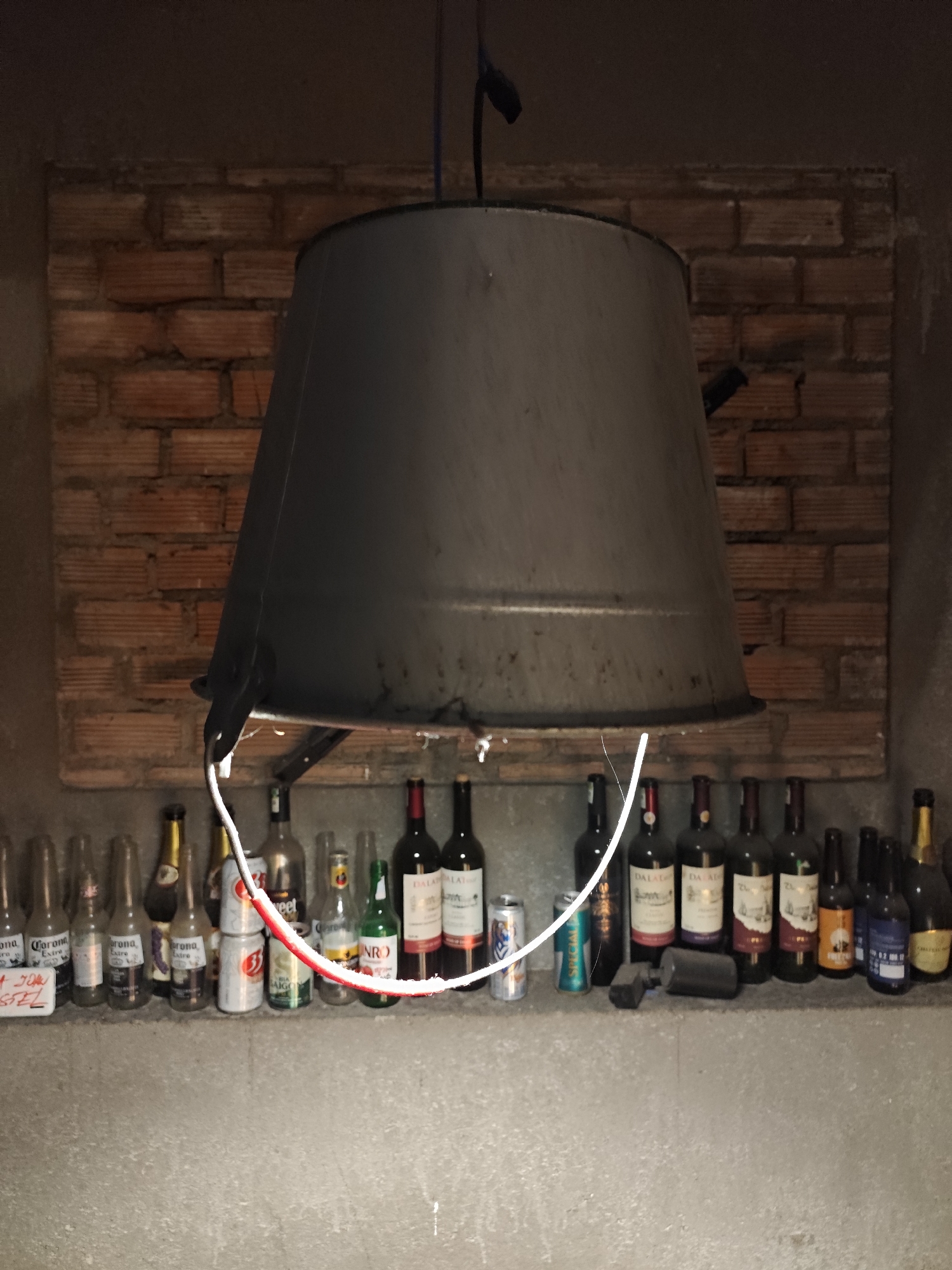 This is an interior image of a casual establishment in Da Lat, Vietnam. The scene is dimly lit, centered on a concrete shelf laden with various alcoholic beverage containers. Above the shelf, a large, dark gray inverted metal bucket serves as a suspended light fixture. A white electrical wire, with a segment showing exposed red insulation, is wrapped around the bucket's lower rim, providing illumination. The background features a rectangular section of exposed red brick wall, bordered by plain, dark gray plaster.
On the concrete shelf, a collection of bottles and cans is arranged. These include clear glass bottles identified as Corona Extra beer, brown glass bottles of Saigon Beer, other green glass bottles, and multiple red wine bottles, several distinctly labeled "DALAT." Additionally, aluminum beverage cans, including one marked "Saigon Special," are present. A small, dark object lies on the shelf near the center-right. No people, explicit actions, or interactions are visible.