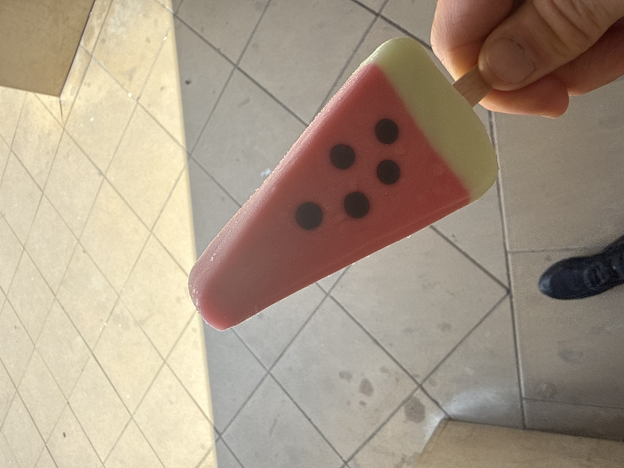 A person's hand holds a watermelon-shaped popsicle against a backdrop of light-colored tile flooring. The popsicle features a red body with black "seeds" and a green rind at the top. The location is outdoors, likely a public area in Sofia, Bulgaria, as indicated by the typical urban tile flooring of the city.