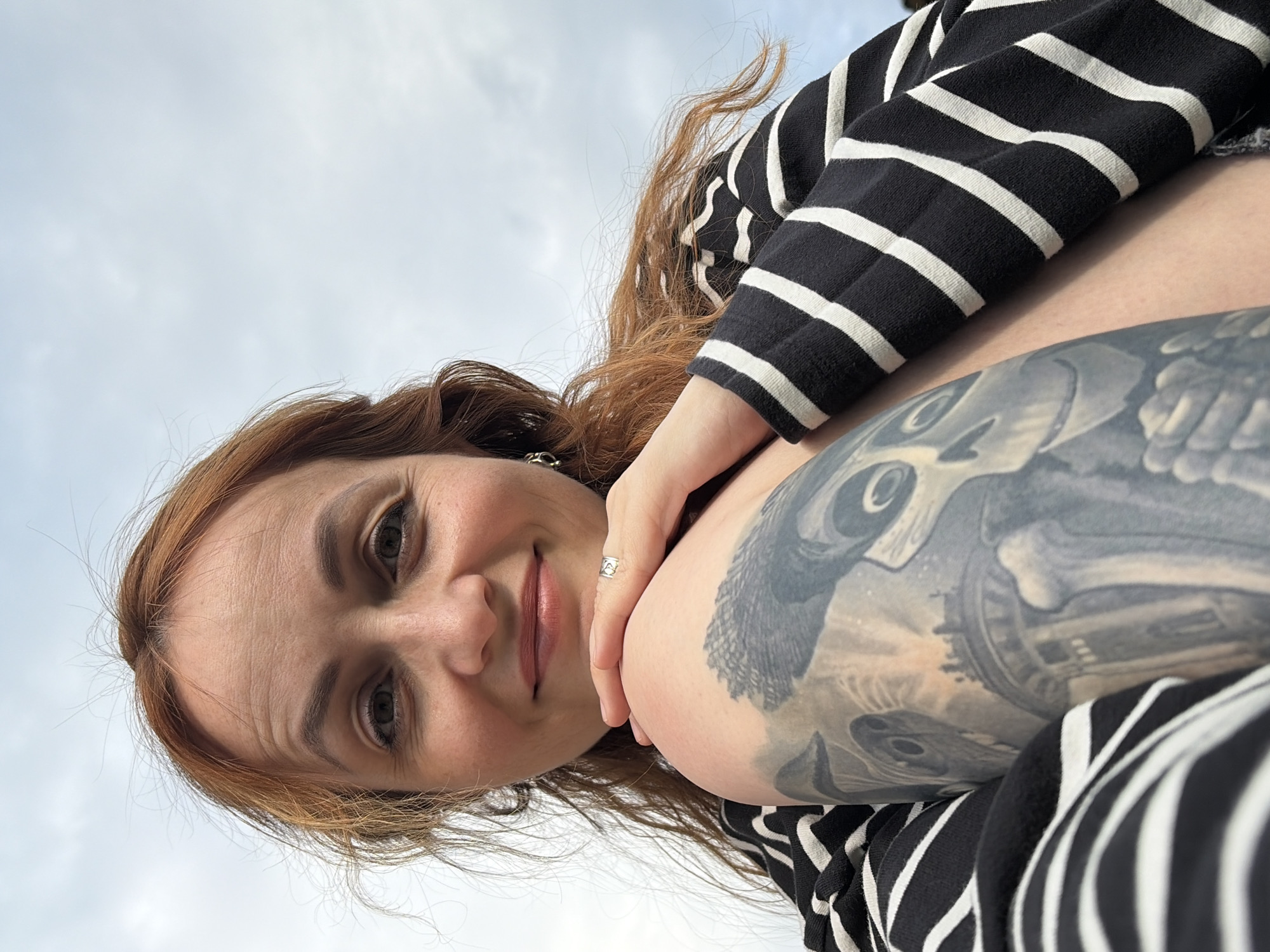 A woman with auburn hair is captured in a close-up, low-angle selfie. She is wearing a black and white striped shirt. Her left arm, adorned with a detailed, grayscale tattoo of an owl, rests on her chest, with her hand gently touching her face. A silver ring with a small heart detail is visible on her ring finger. Her expression is soft and inviting, with a slight smile and her eyes looking towards the camera. The background is a soft, overcast sky, suggesting a daytime setting. There are no visible text elements in the image. The orientation of the image is such that the woman appears to be lying down and looking upwards. The setting appears to be outdoors given the natural light and sky background.