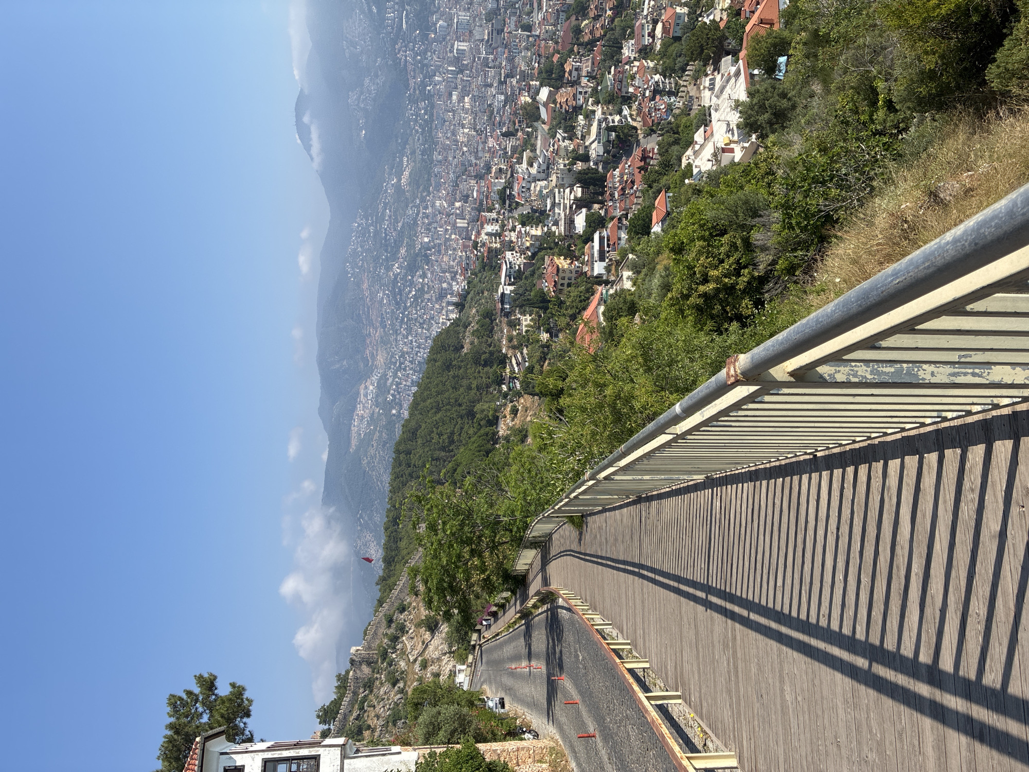 The image shows a scenic view from a pedestrian walkway overlooking the city of Alanya, Turkey. The walkway is constructed of wooden planks and bordered by a metal railing with yellow posts. A cobblestone road runs alongside the walkway, with traffic cones and a few parked vehicles visible. A white building with a red-tiled roof stands on the left, with a large tree growing nearby. The city of Alanya sprawls across the landscape in the background, with densely packed buildings and houses extending towards the hills and mountains. The sky is clear blue, with a few wispy clouds in the distance. A red flag is visible on a distant hilltop, possibly indicating a historical site or a national landmark. The scene conveys a sense of elevated perspective and overlooks a vibrant urban environment nestled within a natural landscape.