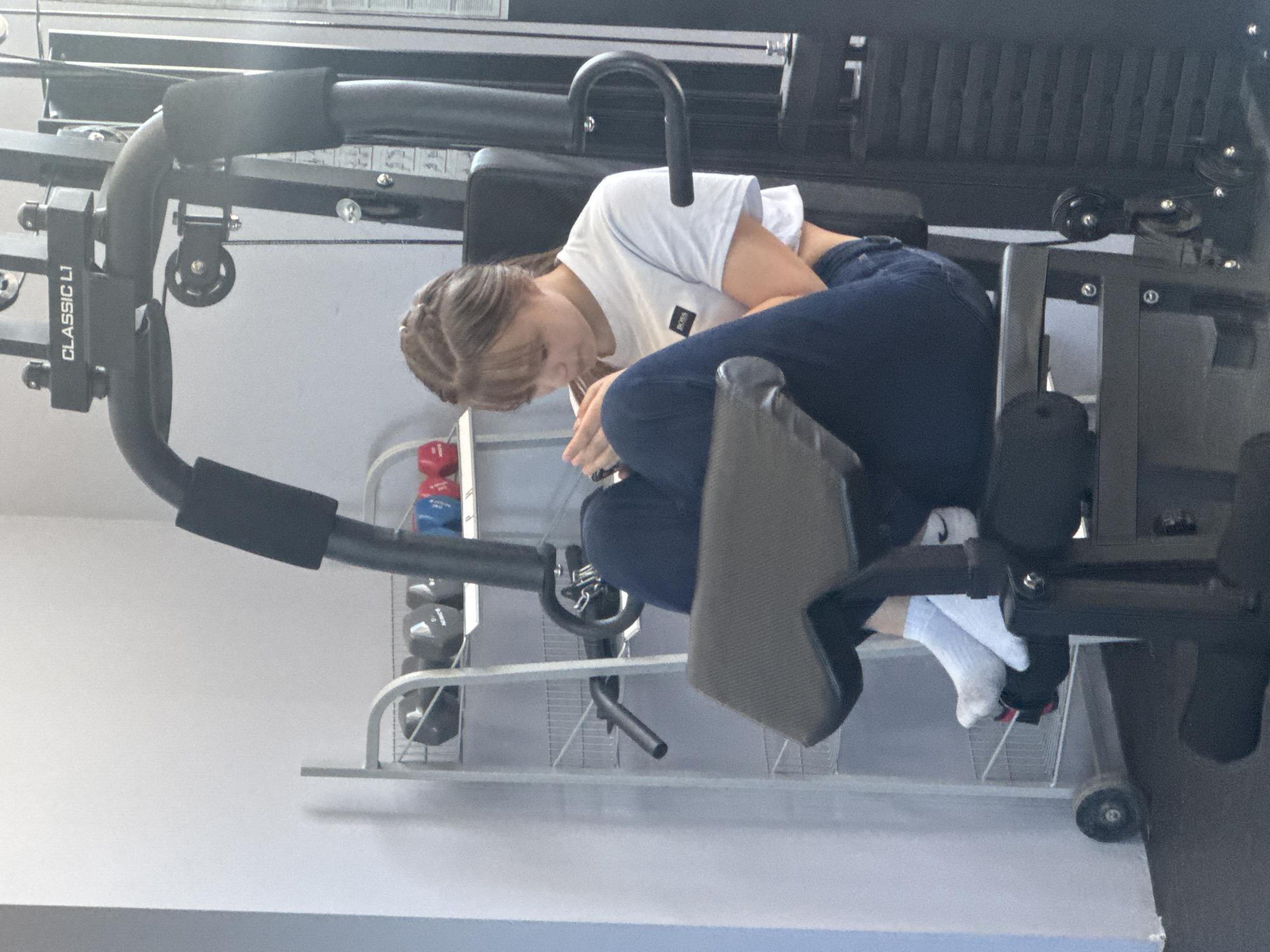The image shows a young woman using a leg extension machine in a gym. She is wearing a white t-shirt and dark jeans, and is seated on the machine with her legs positioned under the padded lever. A rack of dumbbells is visible in the background. The machine has "CLASSIC L1" printed on it. The location is Obruchishte, Bulgaria.