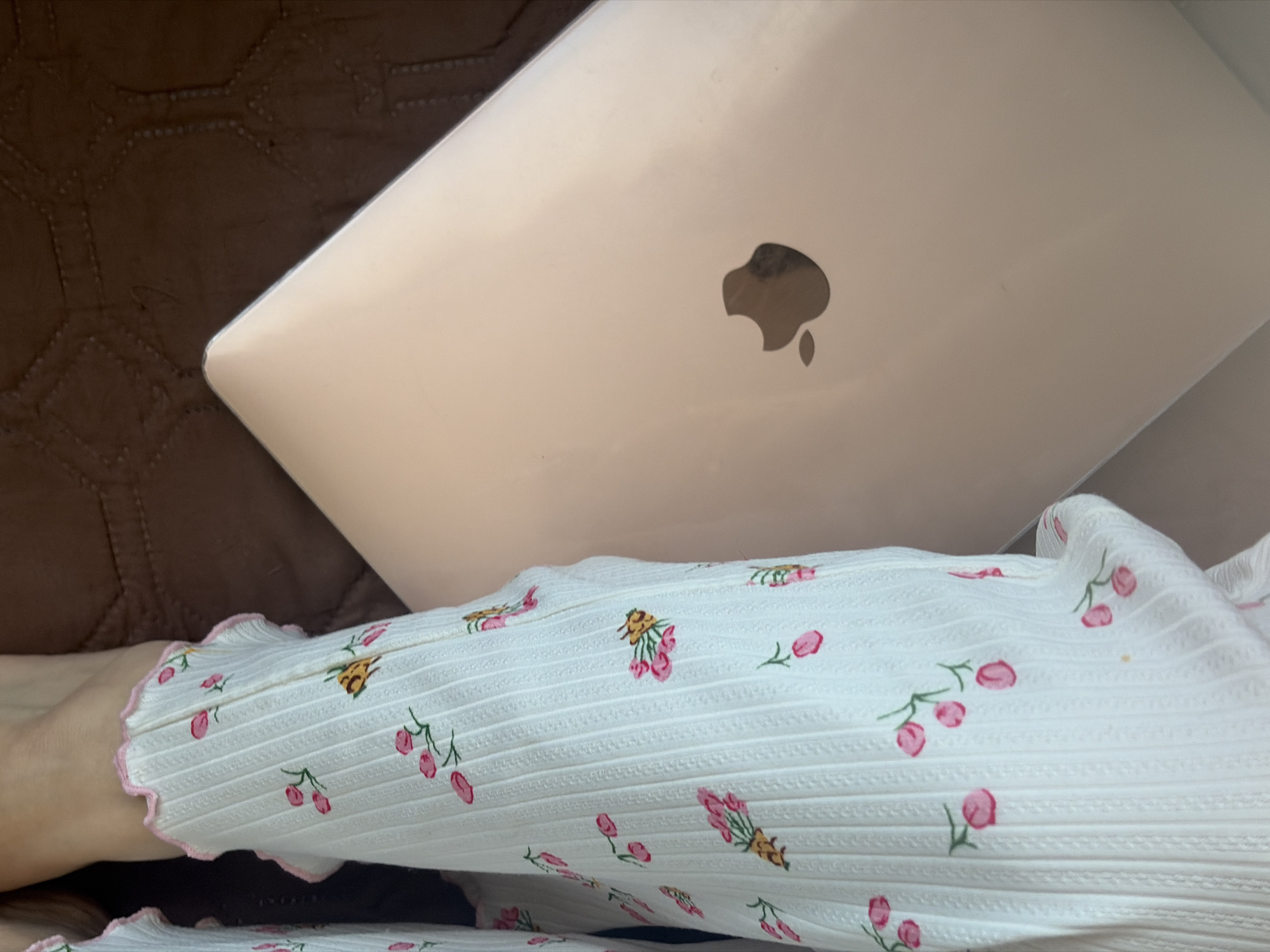 The scene depicts a close-up view of a person wearing a white, ribbed top with a pink floral pattern, seated with a closed Apple laptop resting on their lap. The laptop is light pink. The setting appears to be indoors, possibly on a sofa or bed, indicated by the brown textured material visible in the background.
I am unable to determine the location as Saint Petersburg, Russia with the objects provided.