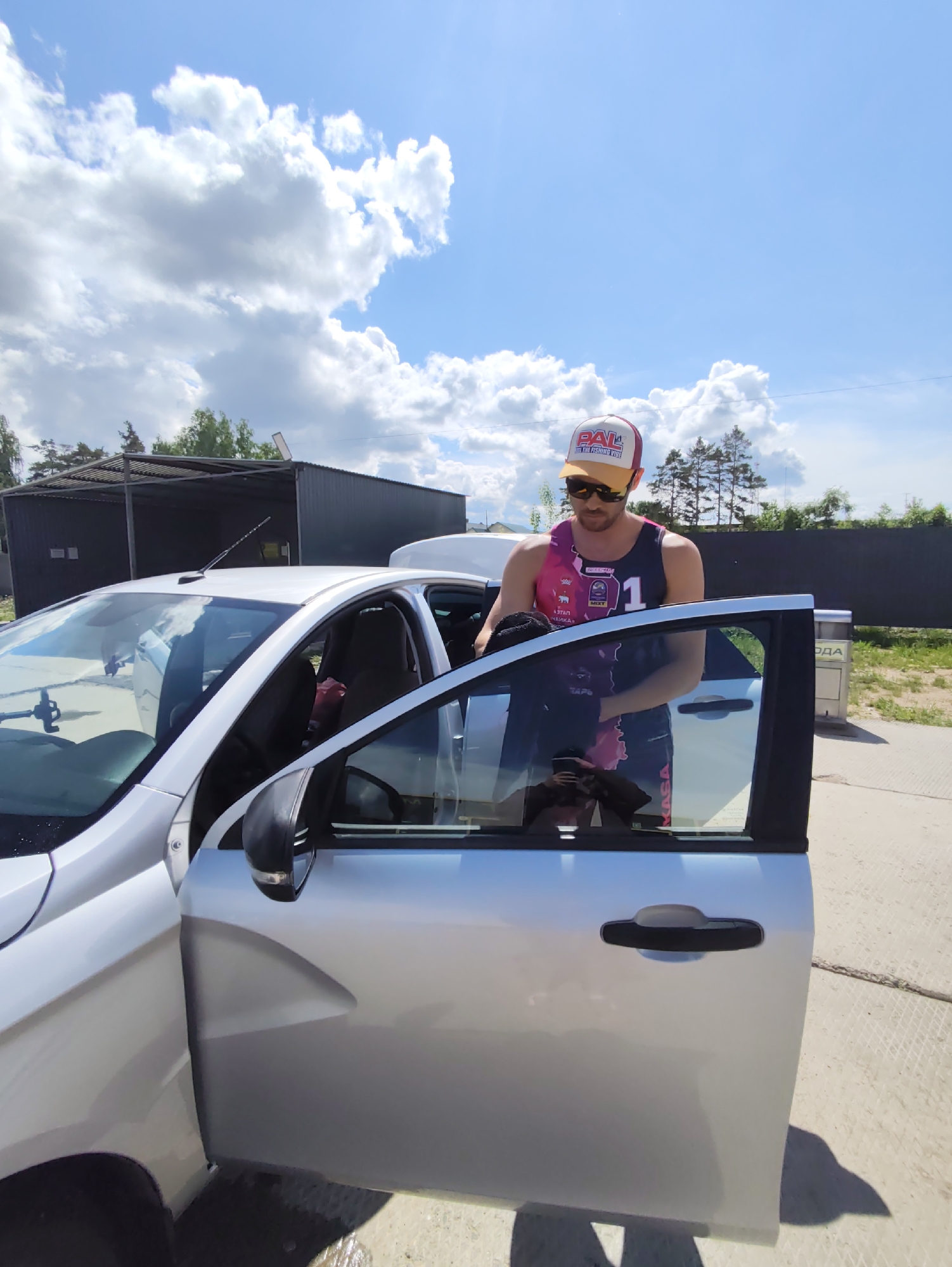 A man wearing a red and white hat, sunglasses, and a pink and black tank top is standing near a silver car with the driver's side door open. He is putting something into the car. The scene is in an outdoor setting on a sunny day with blue sky and scattered clouds. In the background, there is a grey structure and a tree line. Based on the presence of Cyrillic lettering on an object near the car, the location is in Russia, potentially in the city of Chaikovsky.