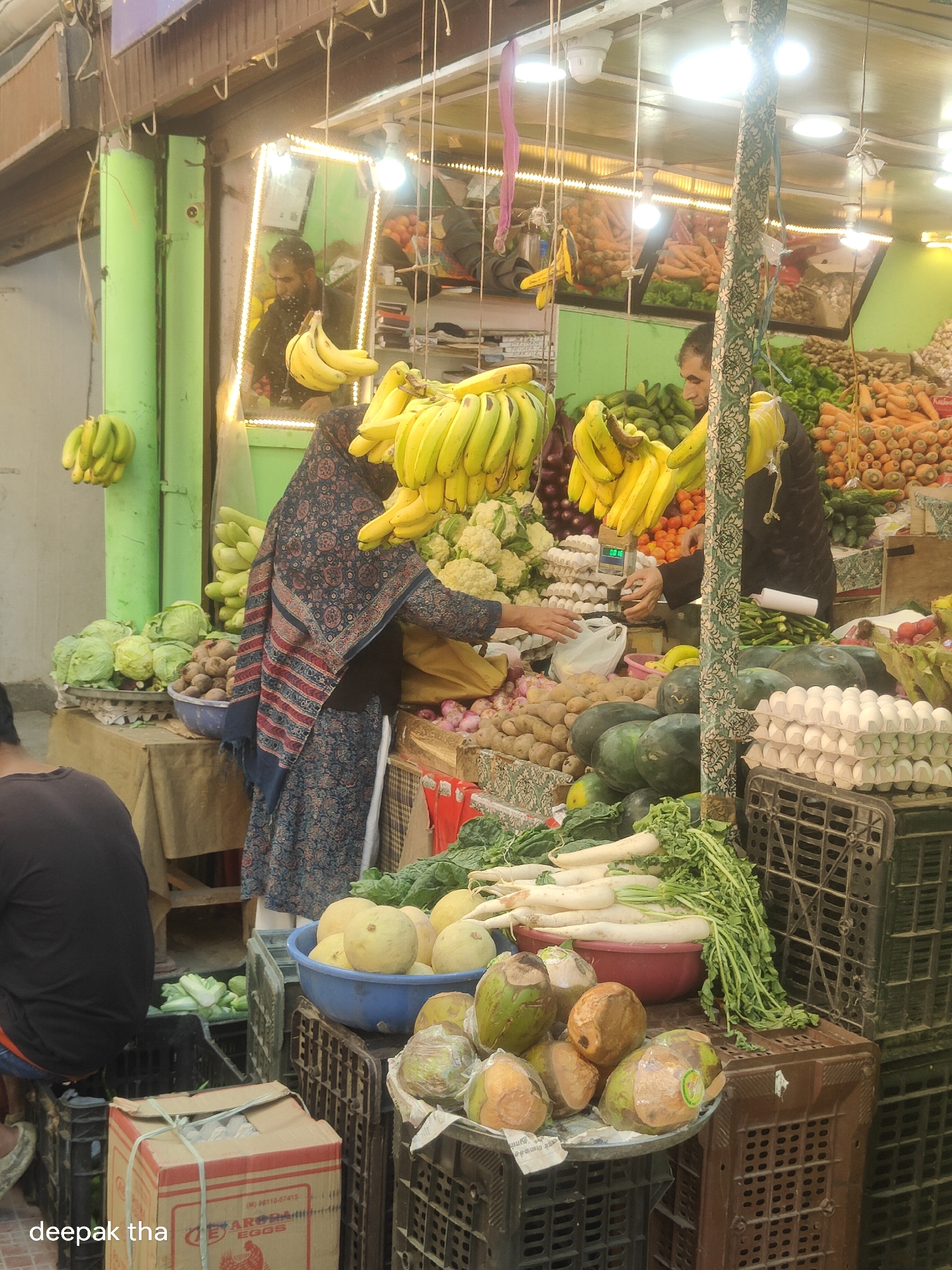 This is a full-shot, eye-level, medium-toned photograph of a bustling market scene, likely in Leh, India, given the presence of the "deepak tha" watermark. The foreground is dominated by a variety of fresh produce, arranged on top of and within black plastic crates. Prominently featured are large bunches of ripe bananas, both yellow and green, hanging from a wooden structure. To the left, a cluster of green bananas hangs from a green pole.
In the mid-ground, a woman in a dark, patterned covering is visible, her face obscured. She appears to be interacting with a vendor or examining produce. To her right, a man, also partially obscured, is handling a plastic bag. Several shelves and displays are laden with a diverse assortment of vegetables, including cauliflowers, cabbages, cucumbers, and what appear to be watermelons. Further back, large quantities of carrots and other root vegetables are visible. In the background, a green wall and various structures suggest the setting is an outdoor market with covered stalls. Several light sources illuminate the scene, casting a bright glow. The overall impression is one of an active marketplace teeming with fresh food and shoppers.