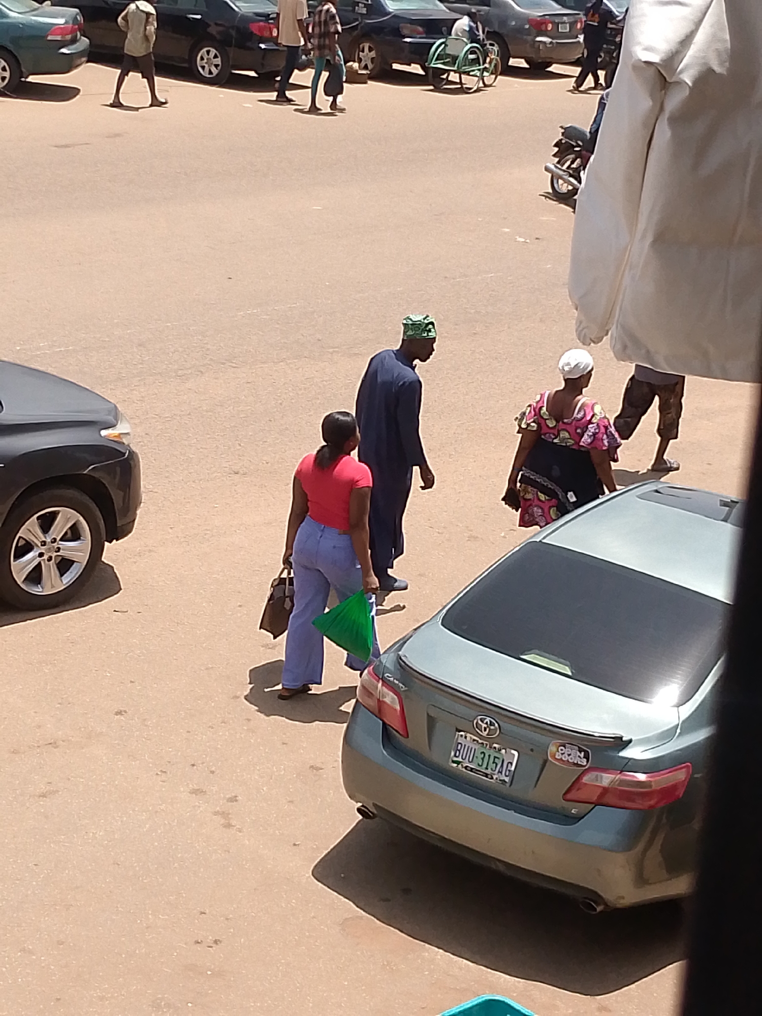 A street scene in Bukuru, Nigeria, shows several people and vehicles on a paved road. In the foreground, a grey sedan is parked, its rear bumper and license plate visible. The license plate reads "BUU 315AG". To the left of the sedan, a dark SUV is partially visible, its front wheel and part of the chassis showing.
Further back, a woman in a red top and blue pants is walking away from the camera, carrying a dark brown bag and a bright green plastic bag. Behind her, a man in a dark blue outfit and a green cap is walking towards the left. Another woman, wearing a colorful patterned dress and a white headscarf, is also walking away from the camera. Several other people and vehicles are visible in the background, including cars and what appears to be a motorcycle. The ground is a dusty, light brown color, and shadows are cast by the vehicles and people. The overall impression is a busy street in a Nigerian city.
