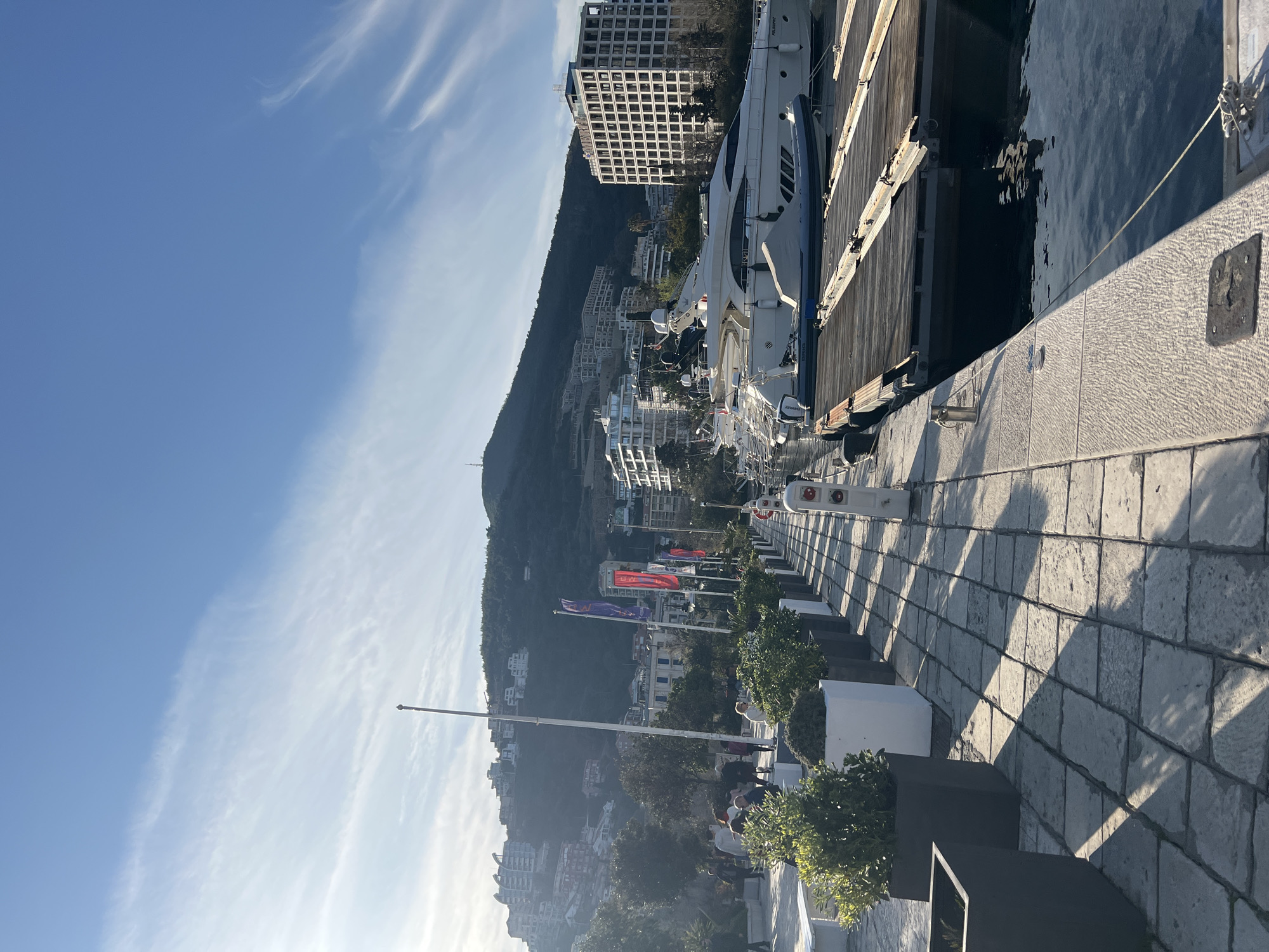 The image depicts a sunlit marina promenade in Budva, Montenegro. The scene is observed from a low angle, looking northeast, with the Adriatic Sea to the right and a steep, developed hillside to the left and background.
In the immediate foreground, a light-colored, roughly cut stone block pavement extends from left to right and recedes into the distance. Long, distinct shadows of planters and unseen objects are cast across this pavement towards the right. Along the left edge of the promenade, a series of square and rectangular planters, some black and some white, hold lush green shrubs. Further along, a few white, minimalist benches are visible.
To the right of the walkway, the dark, calm water of the marina reflects the sky. A thick, light-colored stone quay edge runs parallel to the promenade, with various mooring fixtures, including a silver bollard and a white utility pedestal with red and blue elements, indicating power and water hookups. Beyond this, several wooden floating docks extend into the water.
Moored along these docks are multiple boats, prominently featuring two large white luxury yachts. One yacht, partially covered, has an inflatable dinghy secured to its stern. Another large yacht is marked with "FOLOOSOFIAC" on its stern. Smaller inflatable boats are also visible.
In the mid-ground, a group of indistinct human figures is dispersed along the walkway, suggesting pedestrian activity. Several tall flagpoles, some bare and some displaying vertical banners in red and purple, rise from the promenade.
The background is dominated by a verdant, steep mountain slope dotted with numerous modern, multi-story white and light-colored buildings, indicating extensive residential and commercial development. A prominent, tall, rectangular building with a grid-like facade of many windows stands at the right edge of the image, adjacent to the marina. The sky above is a clear blue with sparse, wispy cirrus clouds, and the strong sunlight suggests either morning or late afternoon.