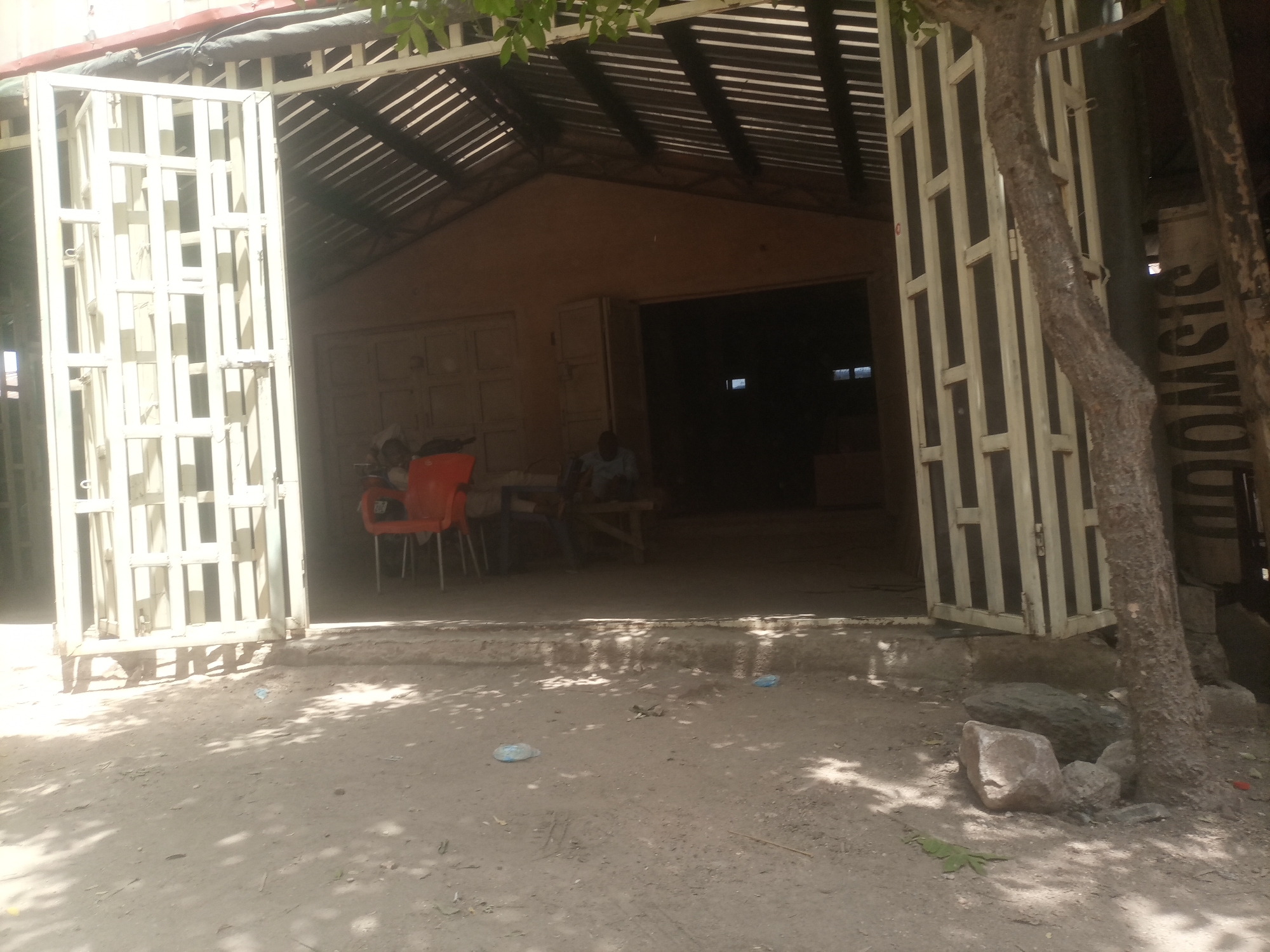 An outdoor scene depicts the entrance to a building, possibly a workshop or shop, located in Arawa, Nigeria. The entrance is partially obstructed by two large, white metal gates, one of which is open. Beyond the gates, the interior of the building is dimly lit, with a dark opening suggesting a doorway or larger passage. A corrugated metal roof supported by visible beams is present above the entrance. In front of the entrance, the ground is covered in dirt and small stones, with dappled sunlight filtering through unseen foliage. Several orange plastic chairs are visible just inside the entrance, and a person is seated on a wooden bench within the building's interior, looking towards the entrance. A rough tree trunk stands to the right of the entrance, and further to the right, a large, vertical sign with dark lettering on a light background is partially visible.