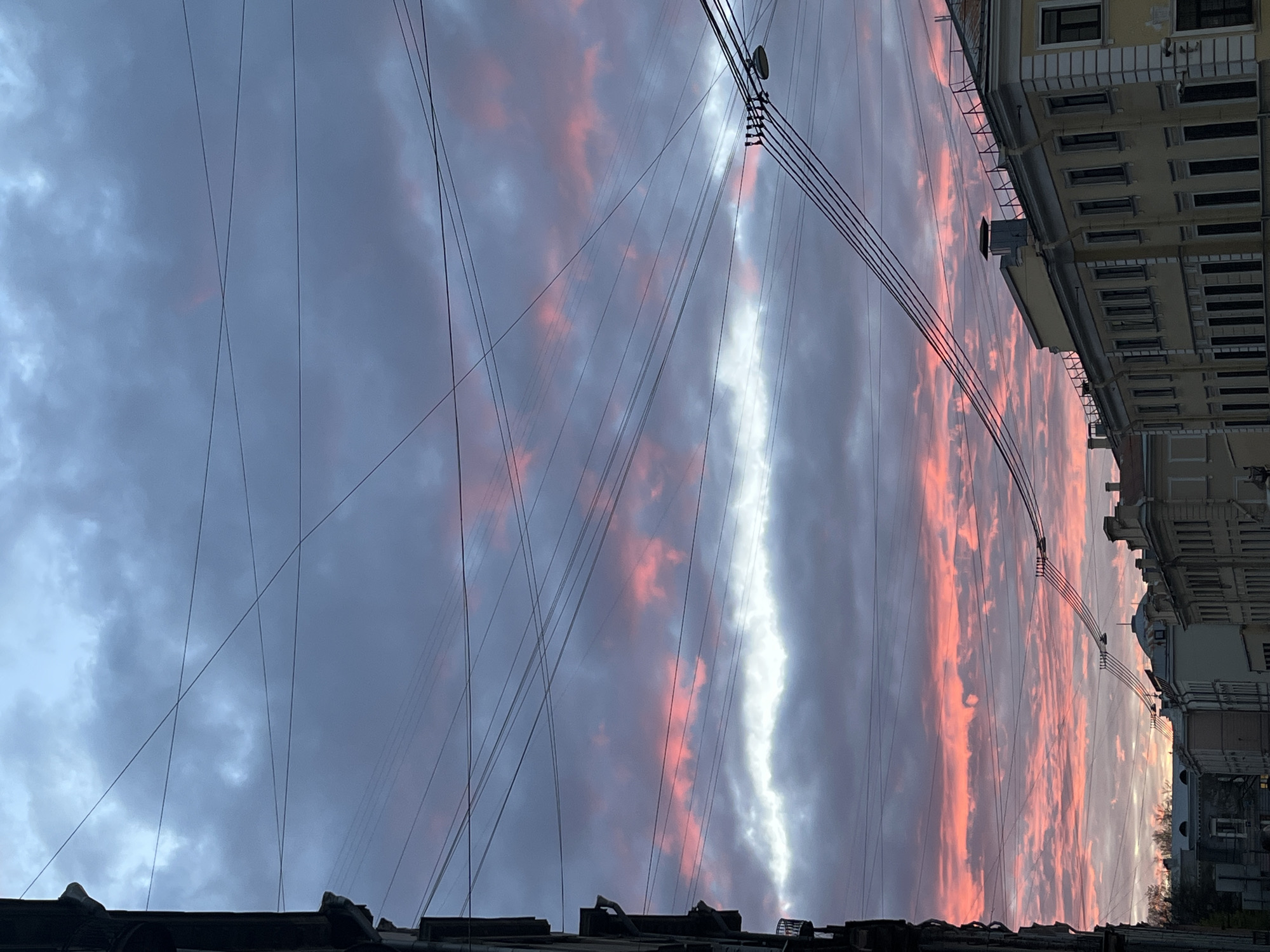 The image is taken from a low vantage point, looking upwards towards a dramatic sky with streaks of pink and grey clouds. Overhead electrical wires crisscross the view. The rooftops of buildings are visible at the bottom of the frame. The sides of the frame include a tan building with windows and ornate facade details characteristic of architecture in Saint Petersburg, Russia. The overall impression is of an urban environment at dusk or dawn, with the sky taking center stage.