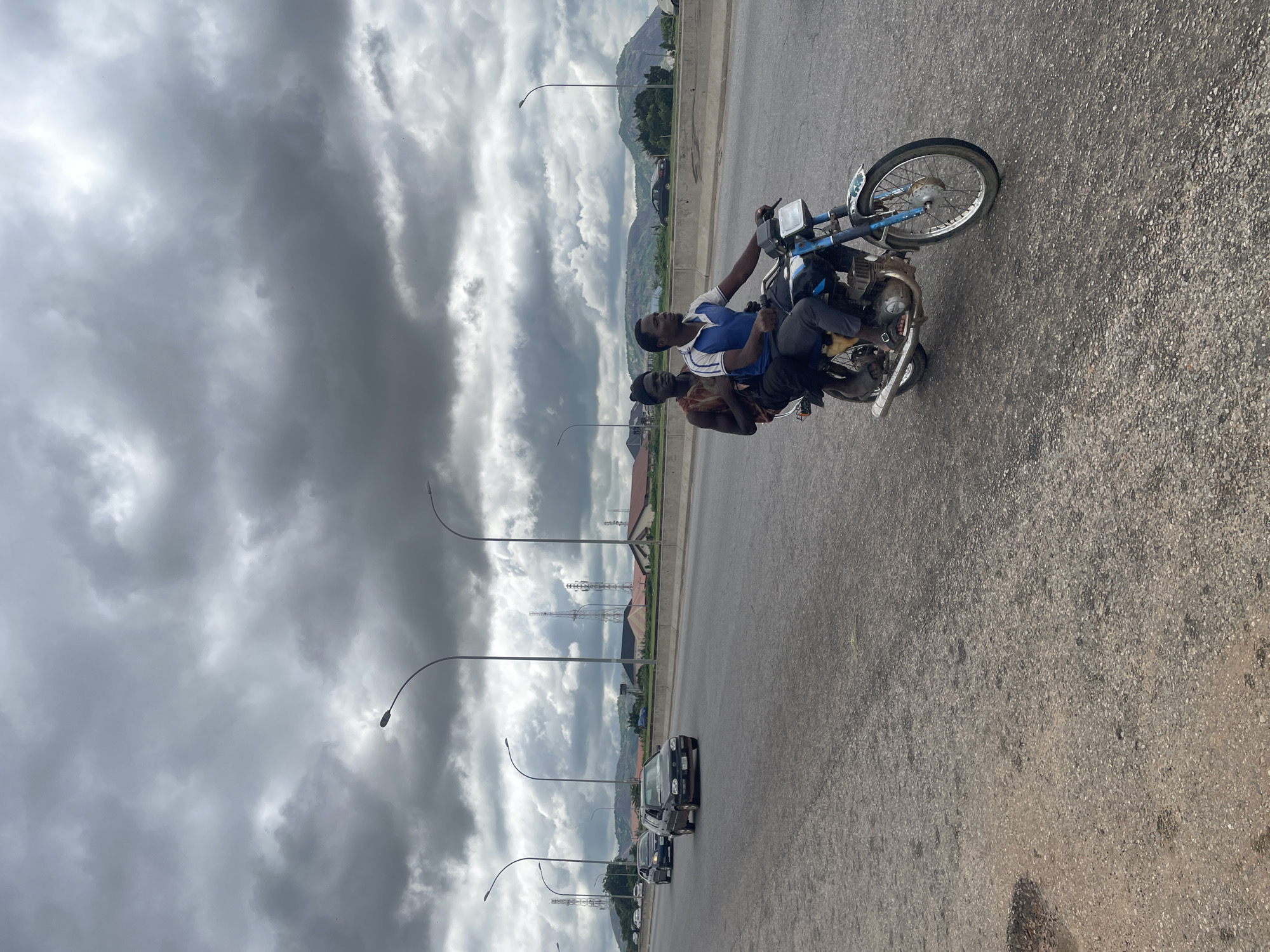 Two young men ride on a motorcycle on a multi-lane highway in Kubwa, Nigeria. The man in front is wearing a blue and white polo shirt and dark pants, while the man behind him wears a sleeveless patterned shirt and dark pants. They are both looking forward. The motorcycle is blue and white with silver accents. The highway is paved and has a concrete divider. In the background, there are streetlights, cell towers, buildings, and rolling hills under a cloudy sky. Several cars are visible on the highway in the distance. The scene appears to be in an urban or semi-urban area.