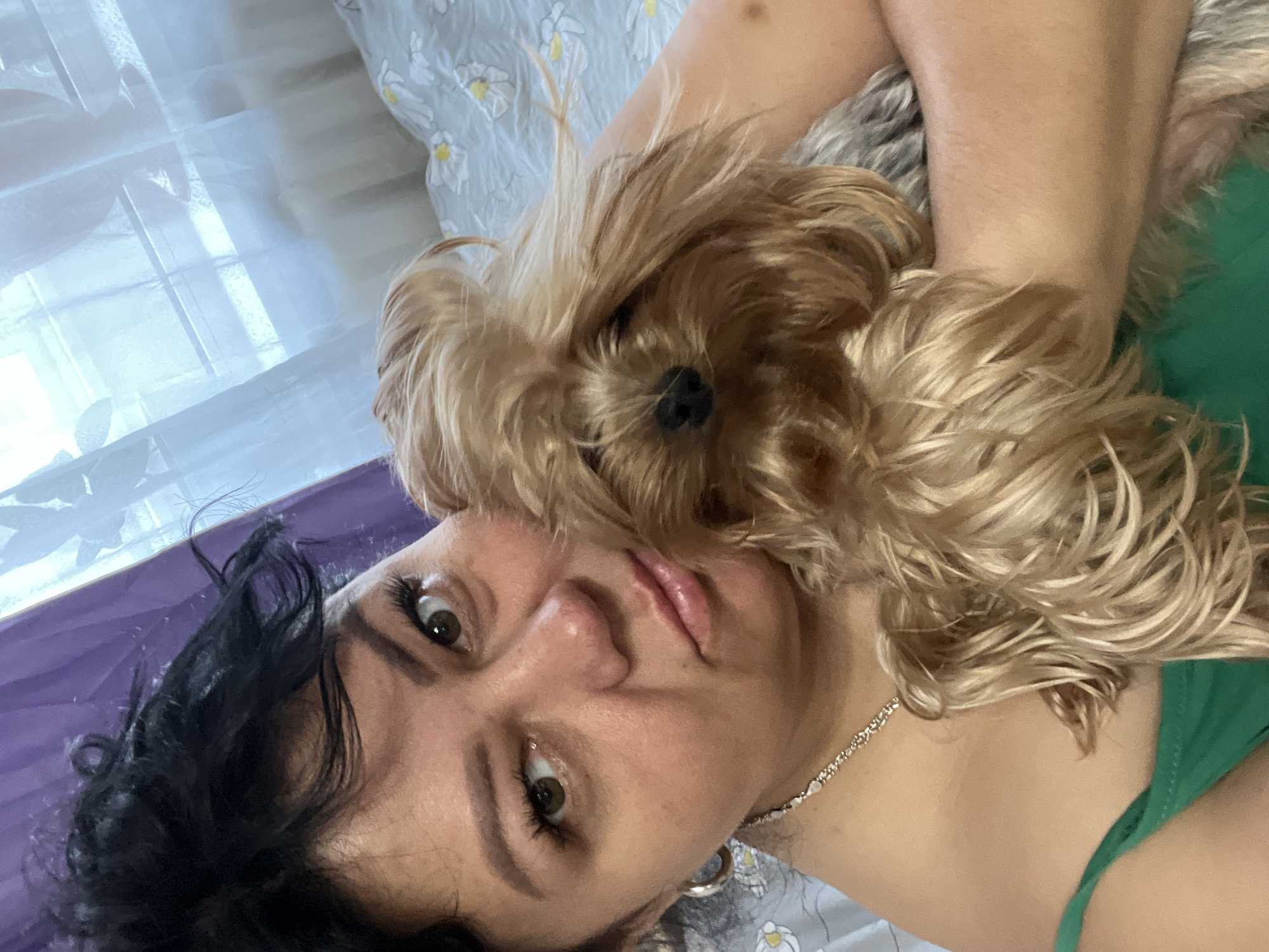 This media file captures a close-up portrait of a woman and her dog, appearing to be taken indoors in Chaikovsky, Russia.
The main subjects are a woman and a small, long-haired dog, likely a Yorkshire Terrier. The woman has dark, curly hair and brown eyes, looking directly at the camera with a neutral or slightly contemplative expression. She wears a silver chain necklace with heart-shaped charms and a green top or garment. Her head is tilted to the left. The dog, with its light brown and blonde shaggy fur, is positioned closely beside her, resting its head near her face, with its dark nose clearly visible.
They appear to be lying down, possibly on a bed or sofa. The background consists of soft furnishings, including a light gray fabric patterned with white and yellow flowers, and a vibrant purple surface behind the woman's head. On the left side of the image, a sheer, light-colored curtain or fabric is visible, suggesting a window and natural light illuminating the scene, indicating it is daytime.
The image depicts a moment of affectionate bonding between the woman and her pet. There is no visible text in the image.