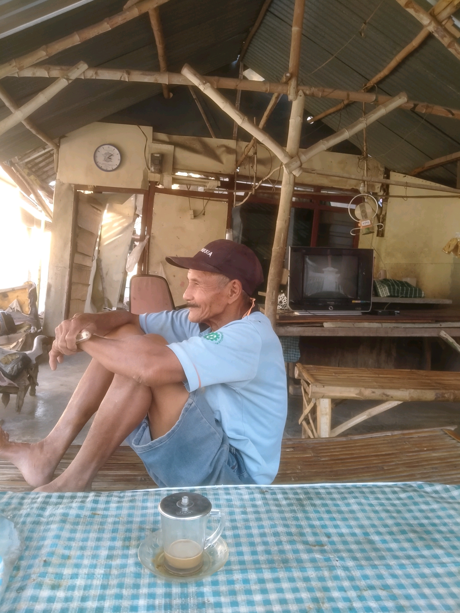 An elderly man, appearing to be of Indonesian descent, sits cross-legged on a woven mat in what appears to be a simple, open-air structure in the city of Sumengko, Indonesia. He wears a light blue short-sleeved shirt and denim shorts, and a dark baseball cap. His hands are clasped in his lap, and he is looking to his right, a slight smile on his face. A glass of coffee sits on a blue and white checkered cloth in the foreground. In the background, a television set sits on a wooden shelf, and other rustic furniture and building materials are visible, suggesting a humble, possibly rural, setting. The structure's roof is made of corrugated metal and supported by bamboo poles. Sunlight streams in from the left, illuminating the scene.