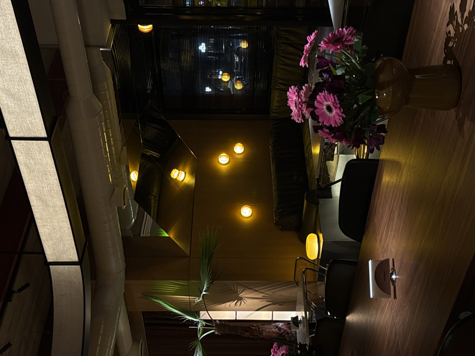 This image depicts the interior of a commercial establishment, specifically Gastrolab, a restaurant/bar located in Yerevan, Armenia. The scene is illuminated by various dim, warm artificial light sources, creating an ambient glow.
In the foreground, a dark wooden communal table occupies the lower portion of the frame. On the right side of this table, a brown ceramic vase holds a bouquet of pink gerbera daisies and other purple flowers. To the left, a small, semicircular, silver-colored napkin holder with a white napkin is placed on the table.
The midground reveals dark-backed dining chairs and an L-shaped dark leather banquette seating arrangement positioned against a smooth, dark mustard-yellow wall. A low, light-colored block-like side table between sections of the banquette features a glowing yellow-orange spherical table lamp. The wall is punctuated by four circular, glowing yellow-orange wall sconces. Above the banquette, a large, angular, dark reflective panel or mirror reflects parts of the interior and the wall lights. To the far left, a tall potted palm tree with large green fronds stands next to dark brown curtains.
To the right of the banquette, a large window fitted with dark horizontal blinds offers a view of a blurred nighttime street scene, showing distant vehicle lights and reflections. The ceiling of the establishment features exposed white ventilation ducts, rectangular overhead light fixtures with translucent fabric diffusers, and black track lighting with individual spotlights. No individuals are visible in the scene.