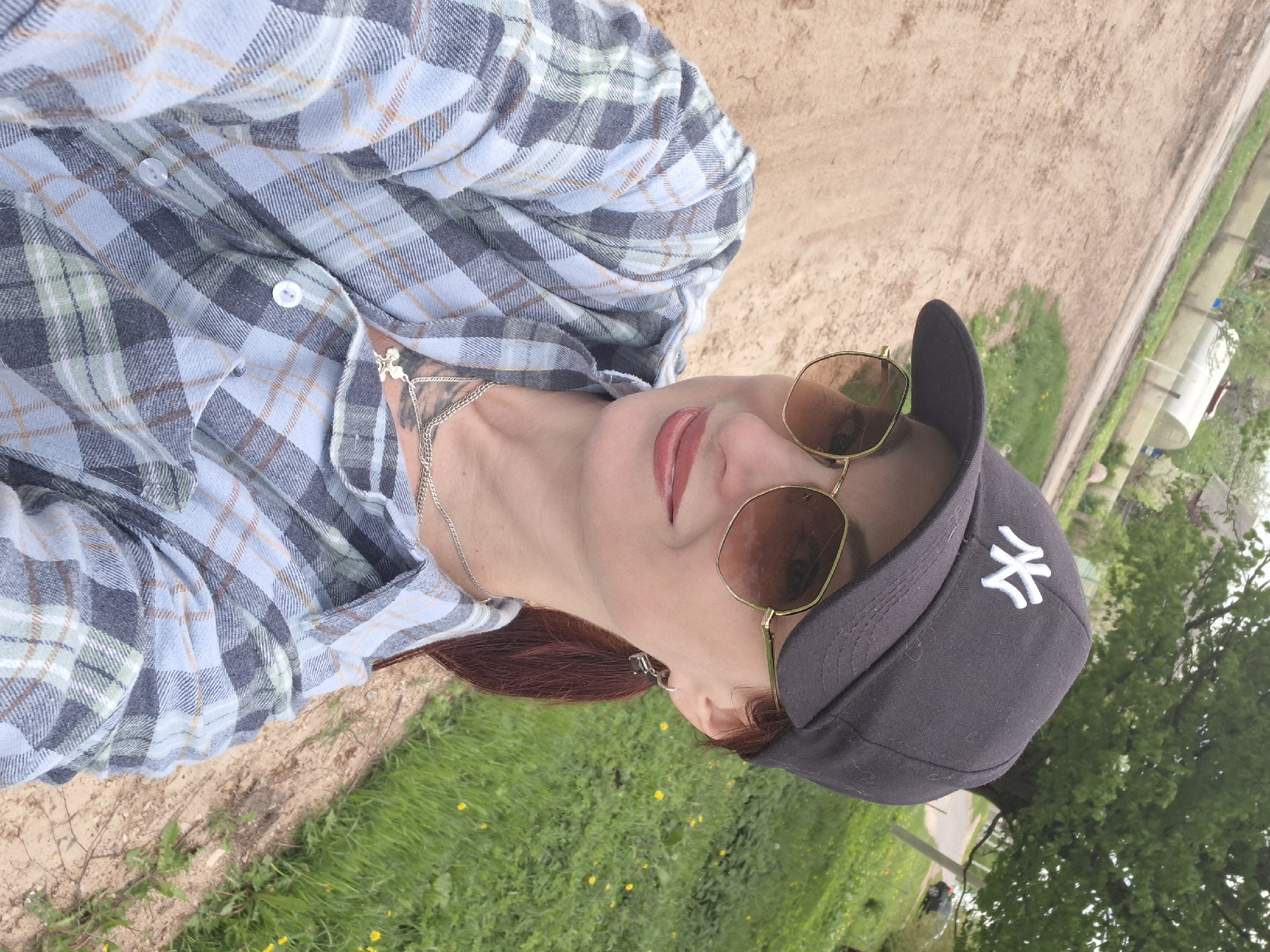A person is pictured from a high angle, wearing a light blue and gray plaid flannel shirt, a silver necklace with a pendant, brown-tinted sunglasses, and a black baseball cap with the New York Yankees logo on it. The person has reddish-brown hair and light skin. The background features a grassy area with small yellow flowers, and a dirt path. Beyond the grassy area, there are trees and possibly buildings in the distance. The location appears to be in a rural or semi-rural area in the city of Забалоцкі сельскі Савет, Belarus.