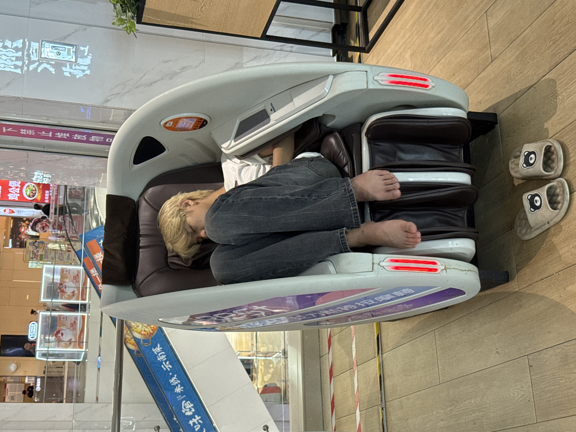 A young person with blond hair is asleep in a white and gray massage chair. They are wearing a white t-shirt and dark gray jeans. Their feet are bare and protrude from the bottom of the chair. The massage chair has brown leather interior and red LED lights on the sides. A pair of beige slippers with bear faces are placed on the light brown wooden floor in front of the chair. The scene appears to be inside a shopping mall in Huizhou, China, with escalators and store displays visible in the background. The floor is made of light brown wooden planks.