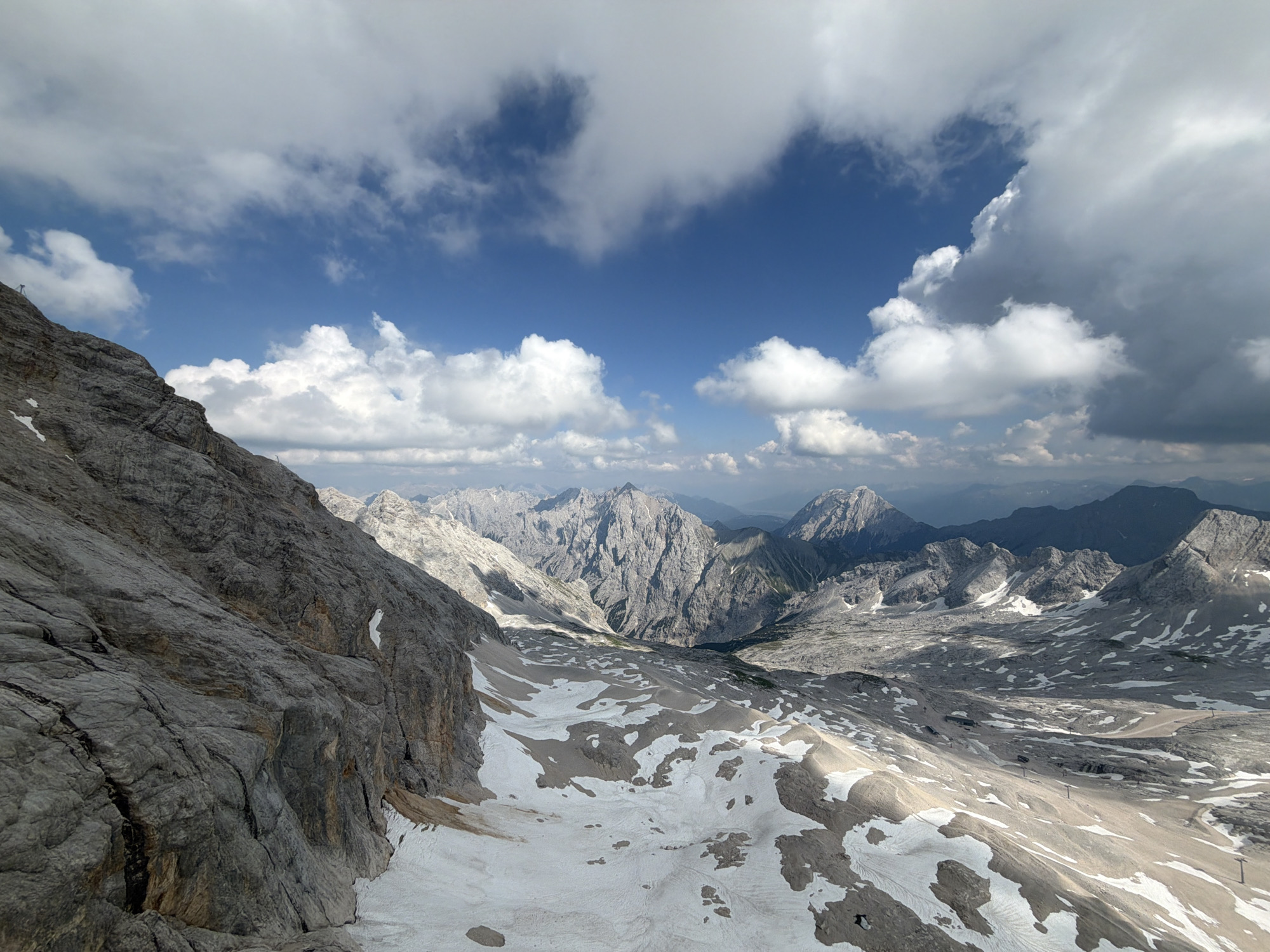 This media file depicts a breathtaking mountainous landscape, likely in the Alps near Garmisch-Partenkirchen, Germany. The scene is a vast expanse of rugged, rocky peaks, some still dusted with snow, indicating a high altitude and possibly early summer or late spring. The foreground is dominated by a steep, textured rock face on the left, showing deep fissures and layers, with patches of snow clinging to its base.
The middle ground opens up to a wide, undulating valley or plateau, characterized by a mix of barren, rocky terrain and scattered snowfields. These snow patches are irregular in shape, resembling a mosaic laid across the lighter-colored rock. In the distance, a dramatic chain of jagged mountains stretches across the horizon, their sharp peaks piercing the sky. The scale of the landscape is immense, conveying a sense of awe and grandeur.
The sky above is a vibrant blue, dramatically contrasted by voluminous white and grey clouds. Some clouds are wispy and streaked, while others are dense and billowy, casting shadows that add depth and dimension to the mountain scenery. The lighting suggests it is daytime, with the sun intermittently breaking through the clouds to illuminate sections of the landscape, creating a dynamic interplay of light and shadow.
No people or specific objects are clearly discernible in the image, placing the focus entirely on the raw, natural beauty of the alpine environment. The overall mood is one of majestic solitude and the power of nature. There is no visible text in the image.