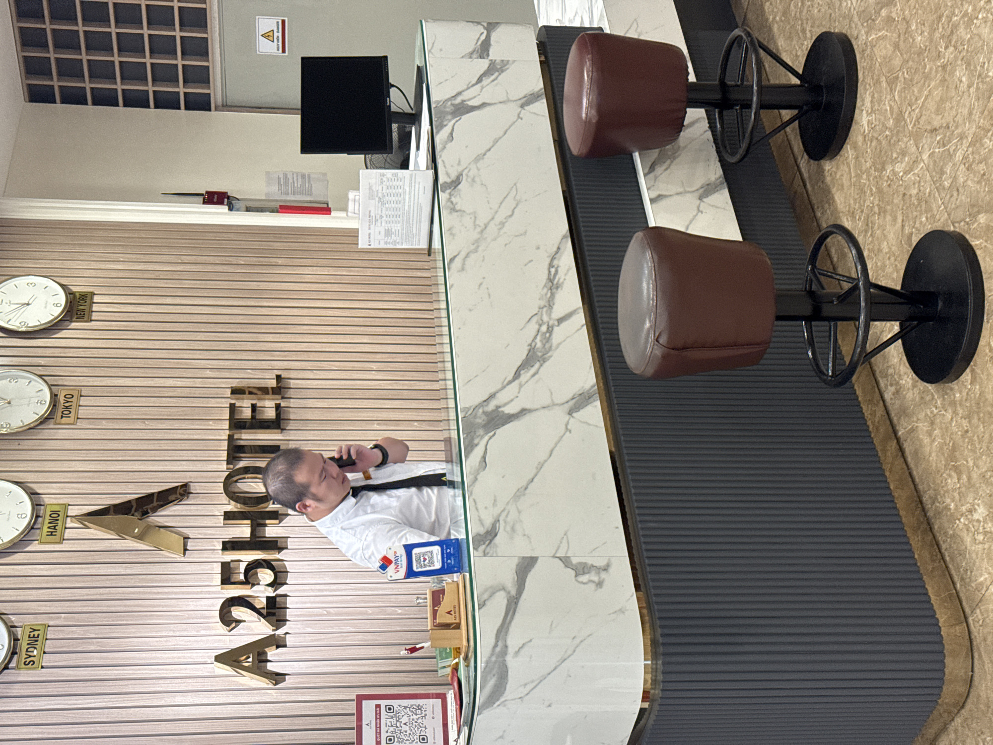 A Vietnamese man in a white shirt and black tie stands behind a marble-topped reception desk, talking on his phone. He appears to be an employee of "A25 HOTEL", as indicated by the prominent signage on the wall behind him, featuring a golden "A" logo above the text. The wall itself is paneled with light-colored wood. Above the hotel sign, four clocks display the times in Sydney, Hanoi, Tokyo, and New York. The reception desk is decorated with a marble pattern and has a black ribbed base. Two dark red bar stools with black metal bases are positioned in front of the desk. On the desk, there are various items including a QR code for VNPay, a pen holder with pens, a sign with "A25 HOTEL" and a price list, a computer monitor, and a red fire extinguisher mounted on the wall to the right. The floor is tiled with a light brown marble pattern.