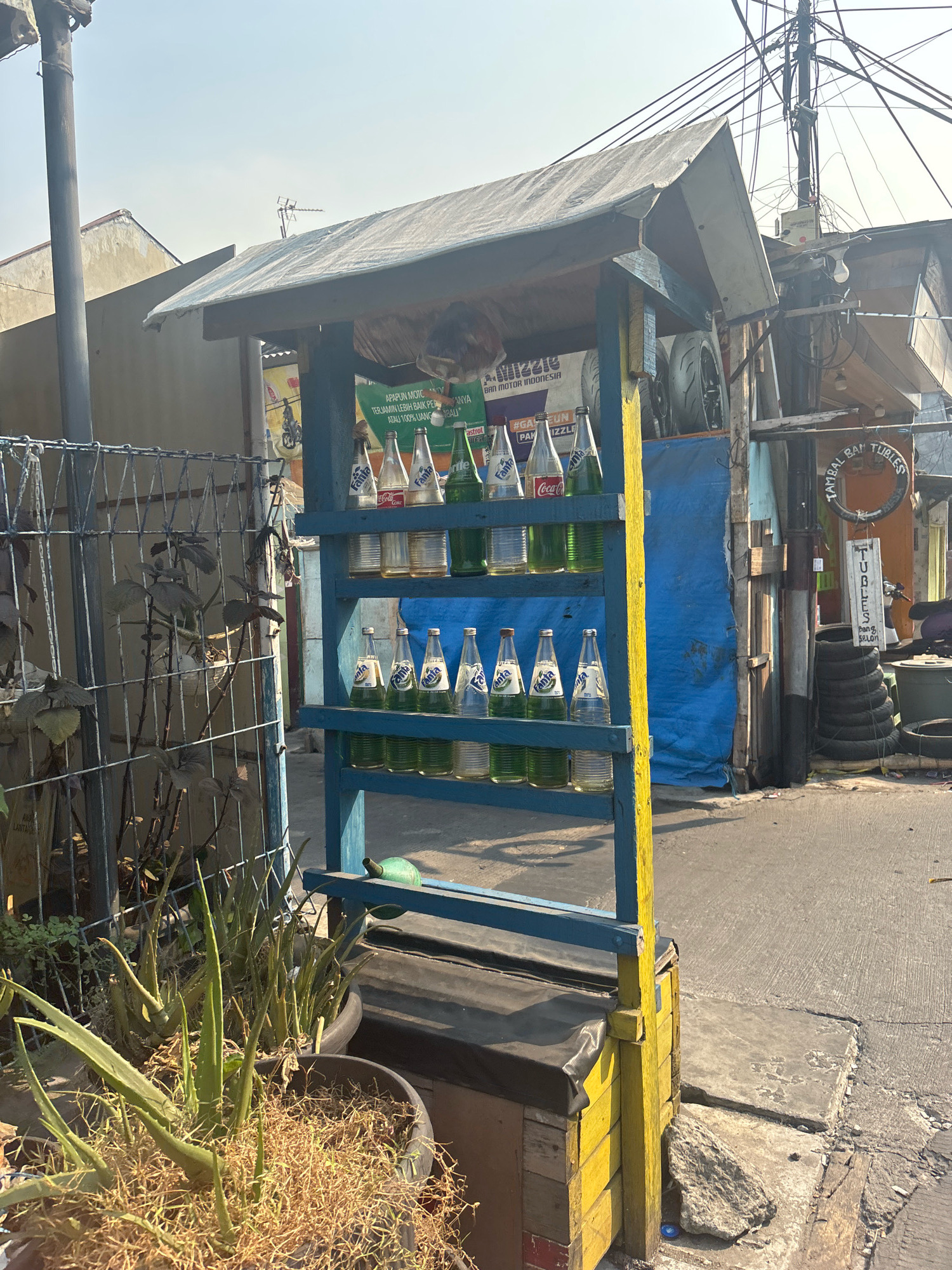 The image depicts a street-side stall in North Jakarta, Indonesia, likely operating as a small convenience store or roadside refreshment stand. The stall is constructed with a blue wooden frame supporting shelves, topped by a weathered grey tarp. The shelves display an assortment of bottled beverages, including Fanta and Coca-Cola, along with several green glass bottles likely containing other soft drinks. In front of the stall, potted aloe vera plants are visible, some with dry, dead foliage.
The environment is characteristic of a bustling urban setting. A concrete sidewalk or road is present, with a wire fence to the left enclosing vegetation. Overhead, a dense network of electrical wires crisscrosses the sky, a common sight in many Indonesian cities. In the background, various structures, including residential buildings and other informal businesses, are visible. Signs with Indonesian text are present, one with the words "TAMBAL BAN TUBELESS," indicating a tire repair service. Stacked tires are also visible, suggesting a connection to automotive services. The overall impression is of a functional, informal commercial operation integrated into the urban fabric of North Jakarta.