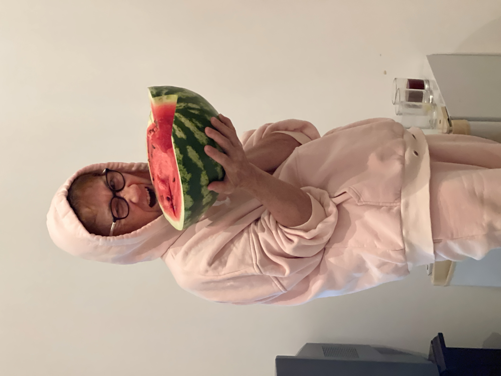 A person wearing a pink hooded tracksuit is holding a half of a watermelon and appears to be biting into it. The person has on glasses and is looking at the watermelon. The background is a plain white wall. There is a television to the left of the frame and a white cabinet with three glasses on it to the right. The scene appears to be indoors, in a domestic setting, possibly in Ornek, Kyrgyzstan.